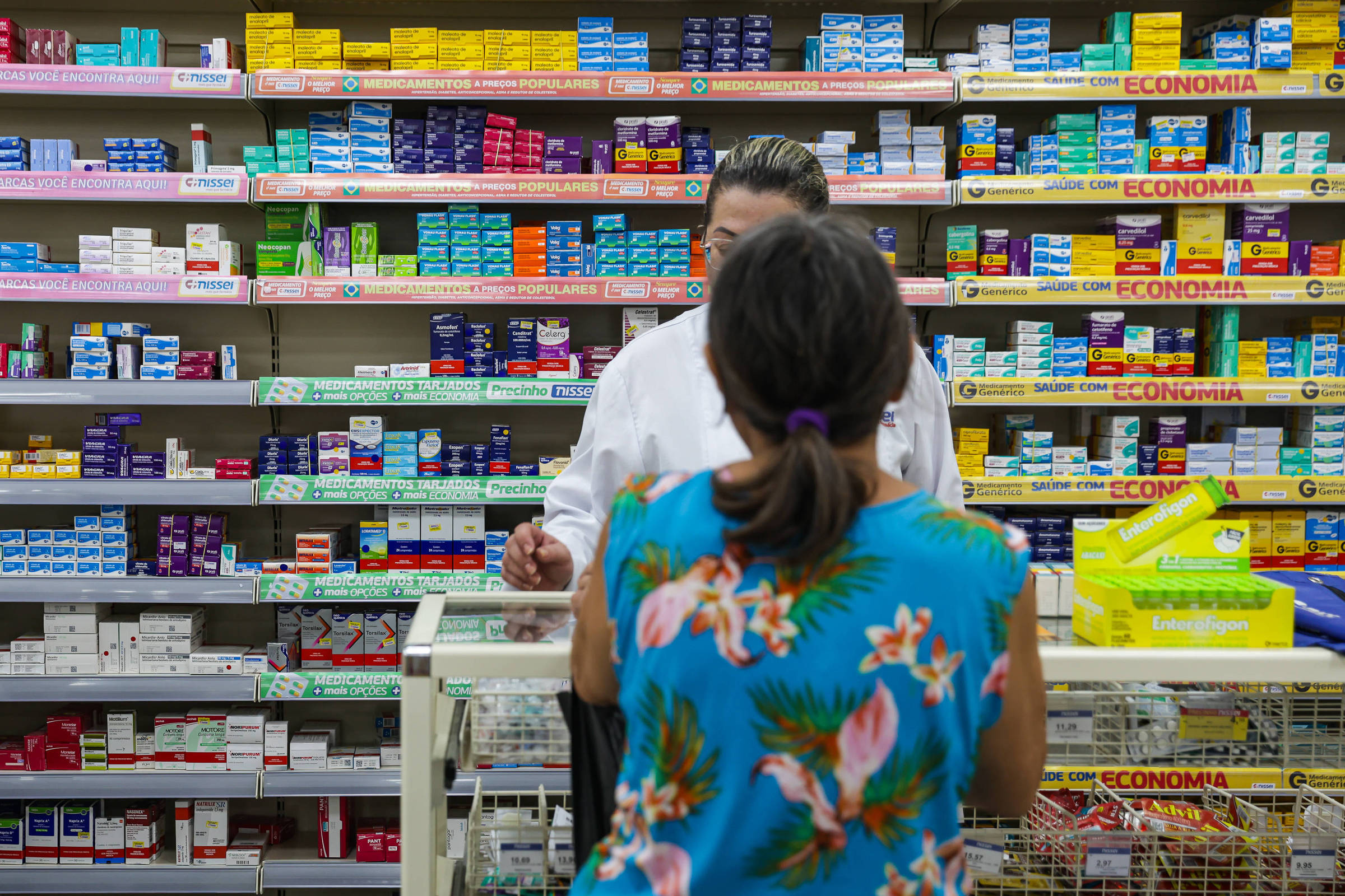 A imagem mostra o interior de uma farmácia, com prateleiras cheias de caixas de medicamentos de várias cores. Um farmacêutico, vestido com um jaleco branco, está atendendo uma cliente que está de costas, usando uma blusa azul com estampas florais. O ambiente é bem iluminado e organizado, com produtos dispostos de forma acessível.