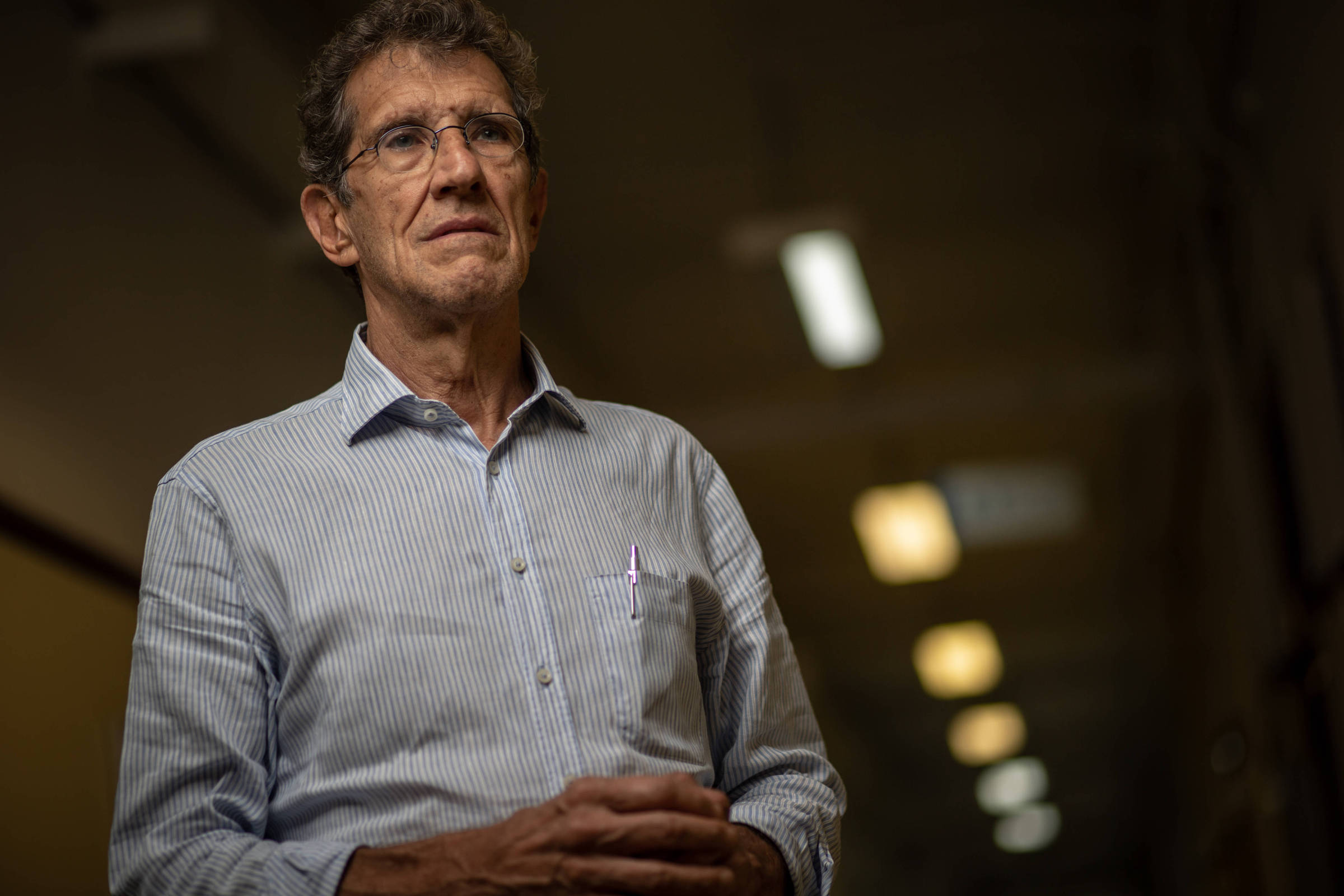 Um homem de cabelos grisalhos e óculos está em um ambiente interno, com iluminação suave ao fundo. Ele usa uma camisa de listras azuis e brancas e parece estar pensativo, com as mãos unidas na frente do corpo.