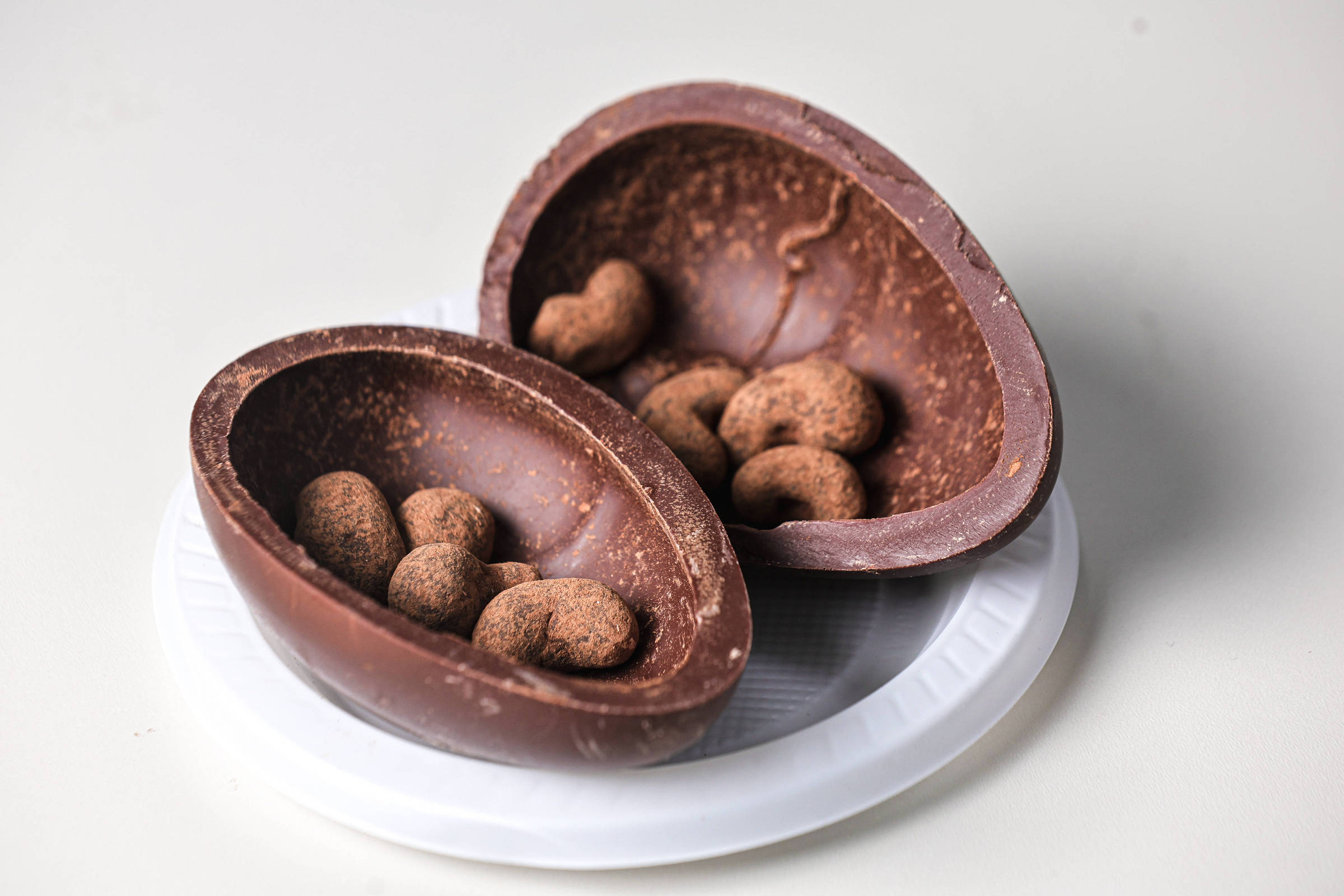 A imagem mostra um ovo de chocolate partido ao meio, colocado sobre um prato descartável. Dentro do ovo, há vários bombons cobertos com chocolate em formato arredondado. O ovo é de chocolate ao leite e apresenta uma textura suave e brilhante.