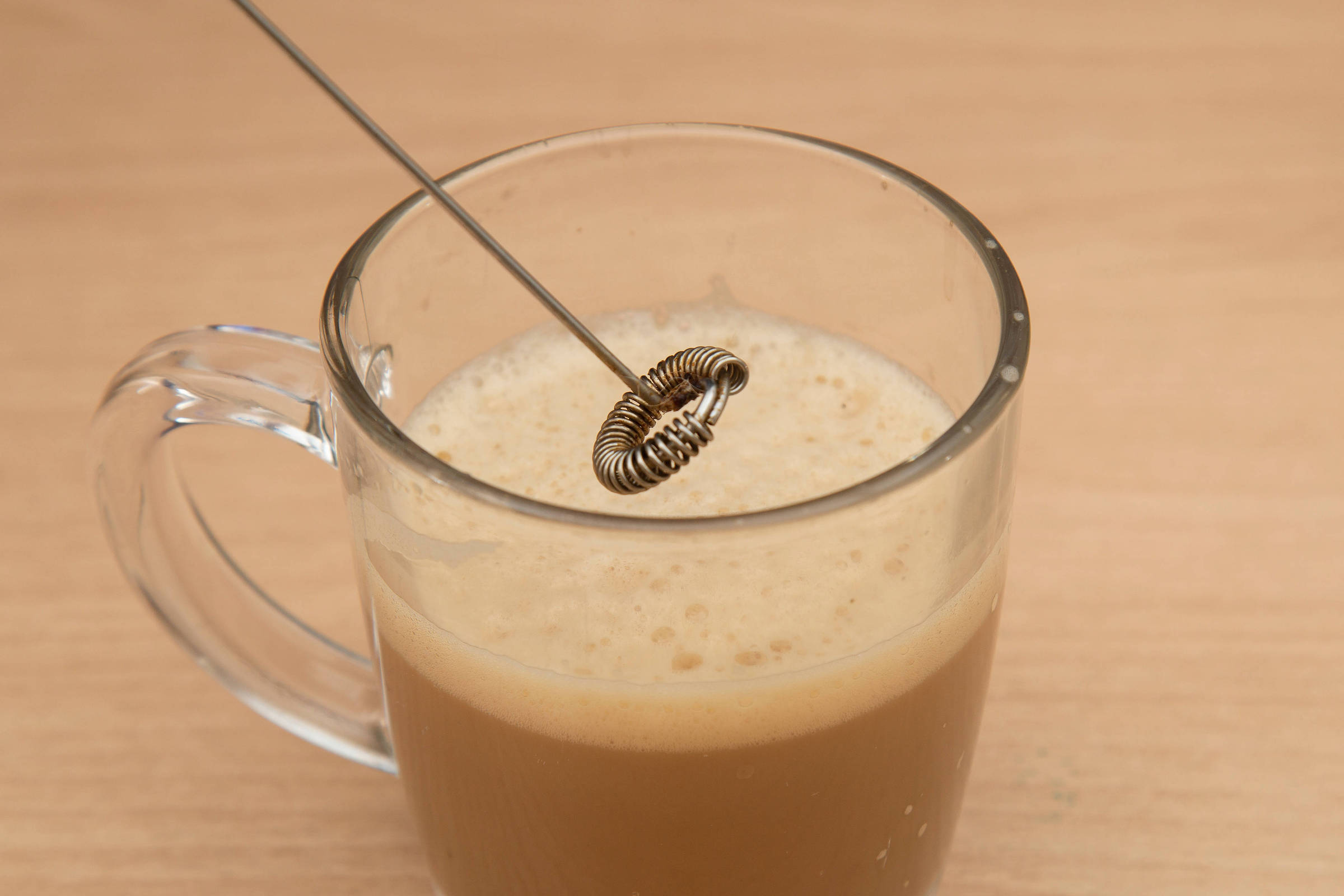 A imagem mostra um copo transparente com uma bebida cremosa e espumosa, possivelmente café. Um batedor de metal, com uma espiral na ponta, está mergulhado na bebida. O fundo é uma superfície de madeira clara.
