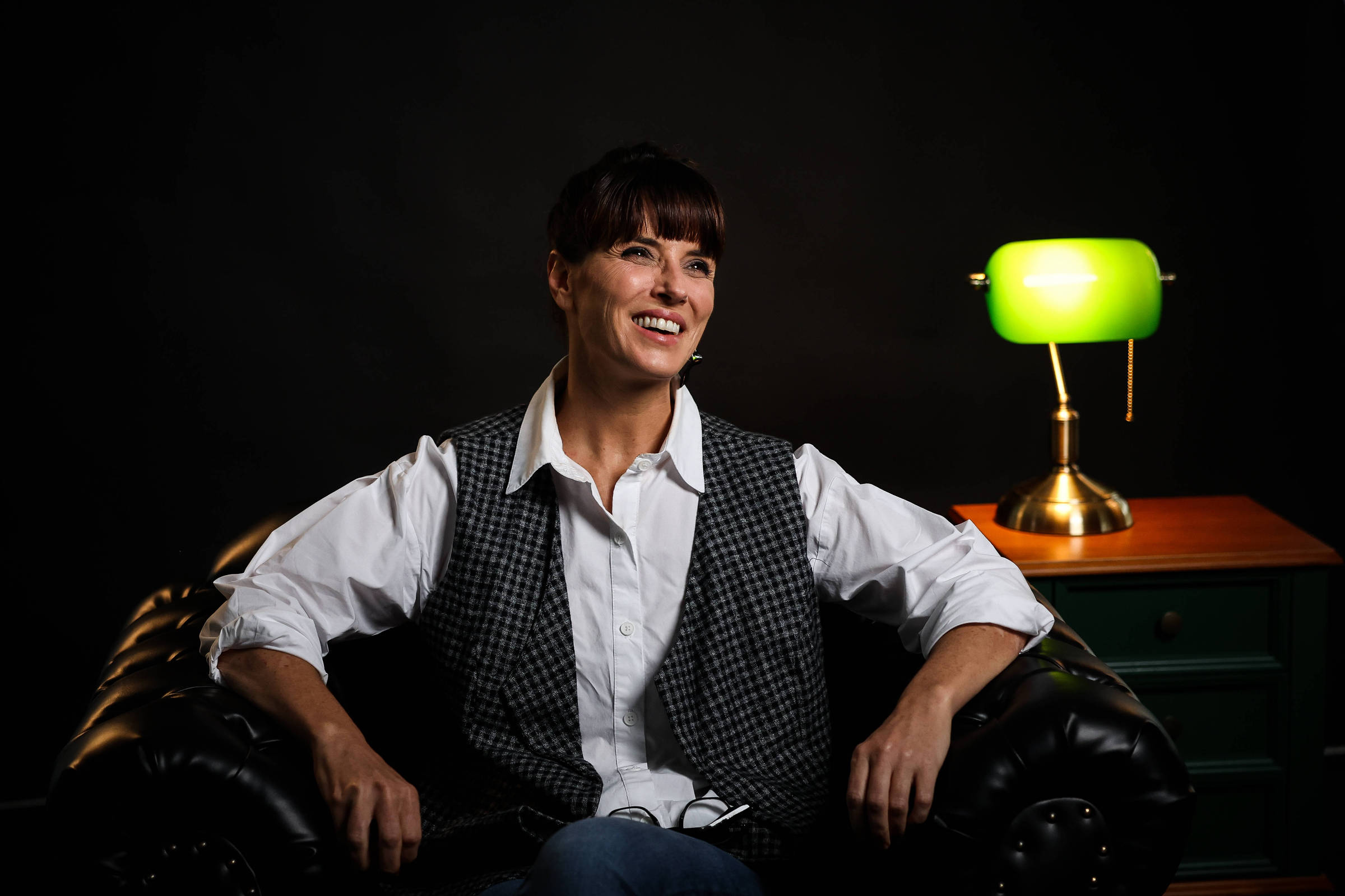 A imagem mostra uma mulher sorridente sentada em um sofá de couro preto. Ela usa uma camisa branca e um colete cinza com padrão xadrez. Ao lado dela, há uma mesa com uma lâmpada verde e uma mesa de cabeceira de madeira. O fundo é escuro, destacando a figura da mulher e a iluminação suave da lâmpada.