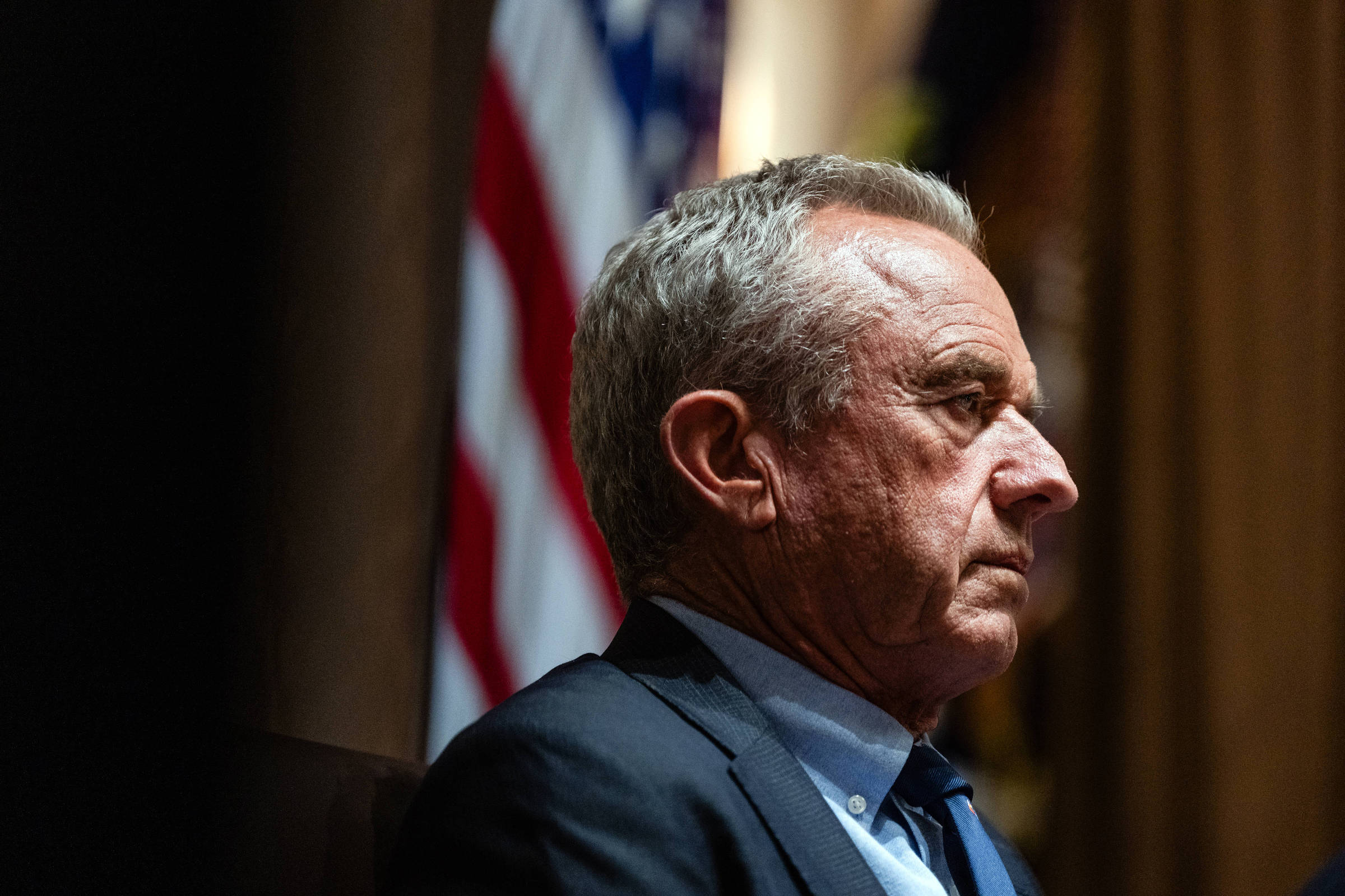 A imagem mostra um homem em perfil, com cabelo grisalho e uma expressão séria. Ele está vestido com um terno escuro e uma camisa clara. Ao fundo, é possível ver uma bandeira dos Estados Unidos, com listras vermelhas e brancas e um campo azul com estrelas. A iluminação é suave, destacando o rosto do homem.