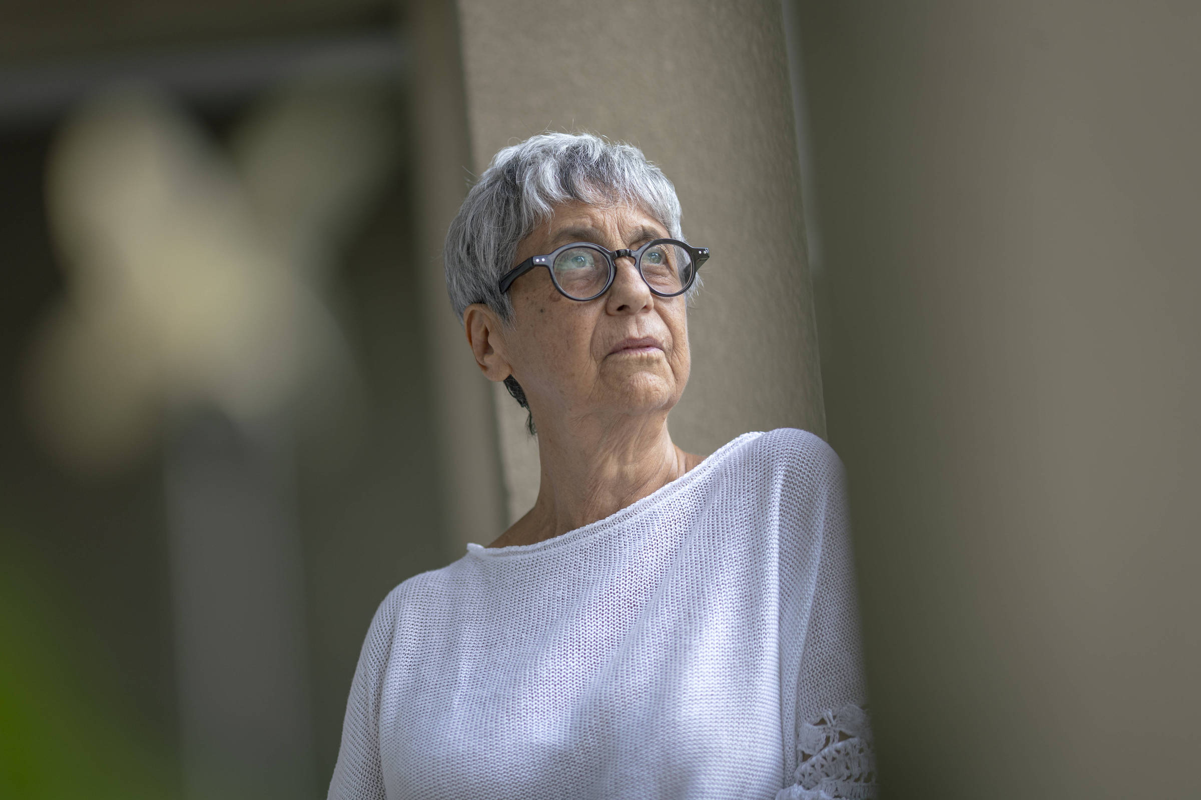 Uma mulher idosa com cabelo curto e grisalho, usando óculos e uma blusa branca de malha. Ela está encostada em uma parede, olhando para o lado, com uma expressão pensativa. O fundo é desfocado, sugerindo um ambiente externo com vegetação.