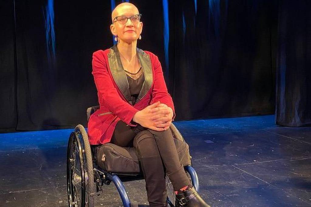 Uma mulher sentada em uma cadeira de rodas, vestindo um blazer vermelho e calças pretas. Ela tem cabelo curto e usa óculos. O fundo é escuro, com iluminação suave, e o chão é de madeira. A mulher está com as mãos sobre os joelhos, olhando para a frente.