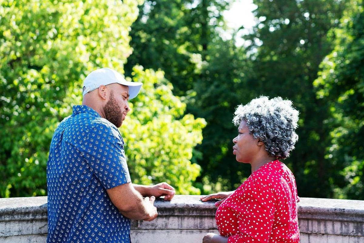 Uma mulher e um homem estão conversando em um ambiente ao ar livre, com árvores verdes ao fundo. O homem usa uma camisa azul com estampas brancas e um boné branco, enquanto a mulher está vestida com uma blusa vermelha com bolinhas brancas e tem cabelo grisalho. Ambos parecem estar envolvidos em uma conversa atenta.