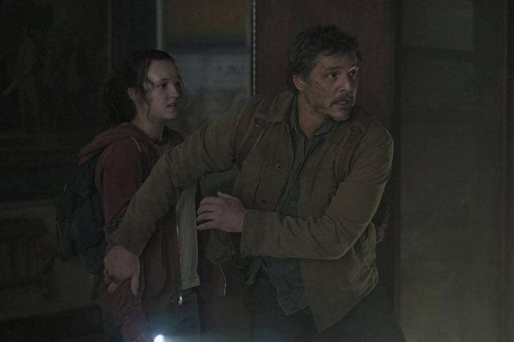 A imagem mostra duas pessoas em um ambiente escuro e abandonado, possivelmente um museu ou galeria. Um homem, com cabelo curto e barba, está segurando uma lanterna e parece estar em alerta, enquanto uma jovem mulher, com cabelo longo e preso, está ao seu lado, segurando sua mão. A iluminação é baixa, criando uma atmosfera tensa e misteriosa.