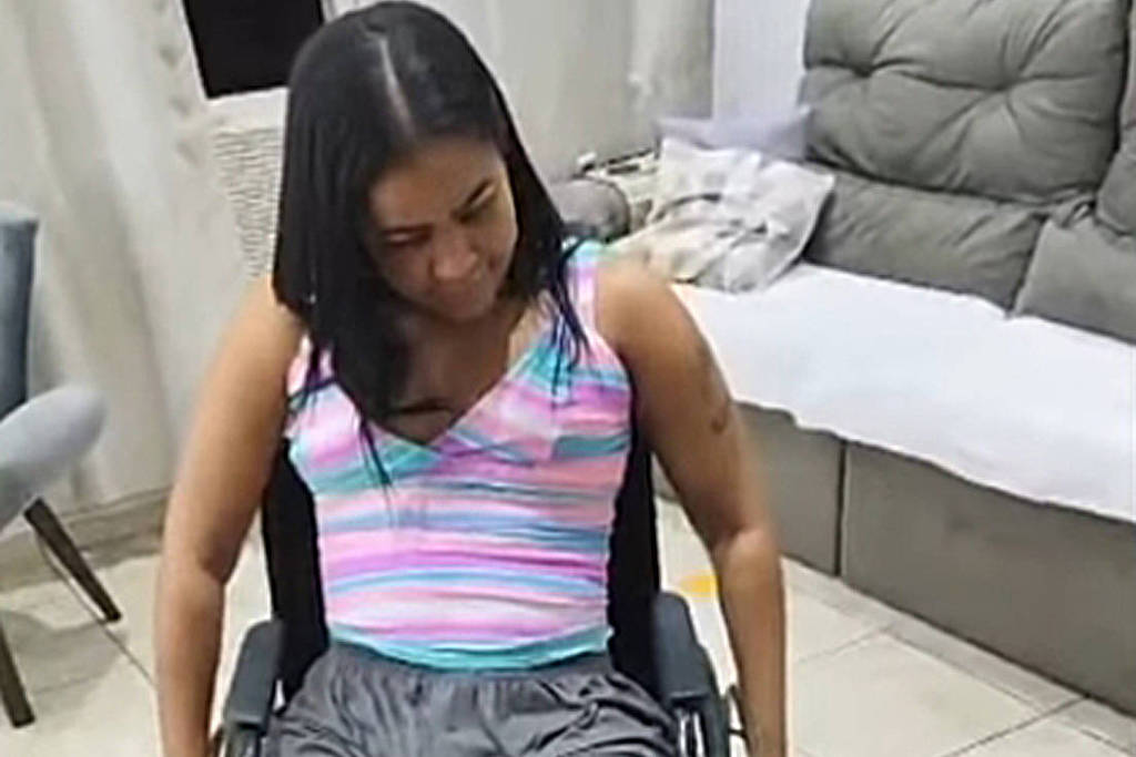 Uma mulher sentada em uma cadeira de rodas, vestindo uma blusa listrada em tons de rosa e azul, e shorts cinzas. Ela está olhando para baixo, com uma expressão de concentração. O ambiente ao redor é uma sala com piso de cerâmica e móveis como uma mesa e um sofá ao fundo.