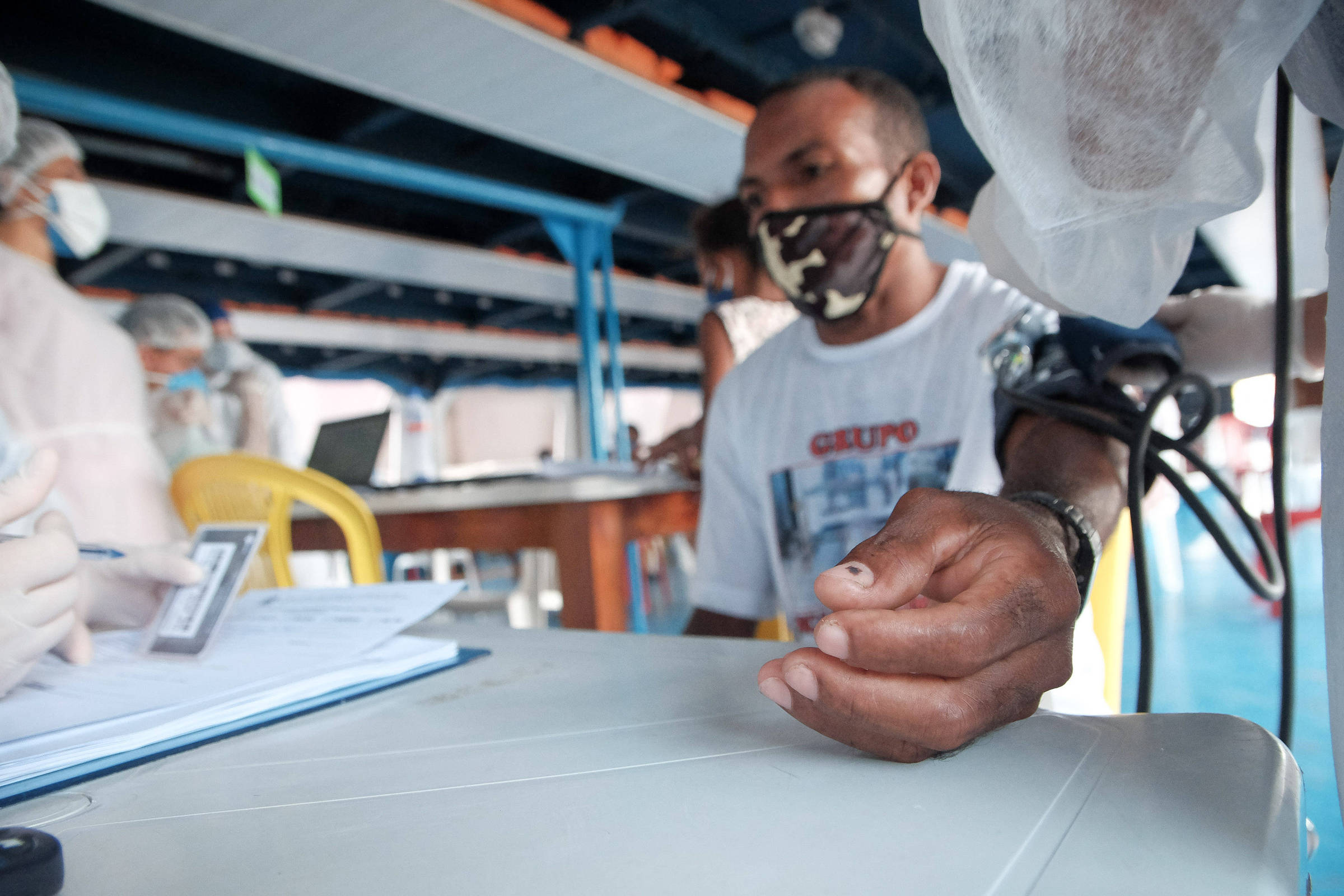 A imagem mostra um homem sentado em uma mesa durante um atendimento de saúde. Ele está usando uma máscara e um colete, enquanto um profissional de saúde mede sua pressão arterial. O ambiente é um espaço interno, com mesas e cadeiras, e outras pessoas ao fundo também usando equipamentos de proteção. O homem tem a mão apoiada na mesa, e há um formulário e uma caneta sobre a mesa.