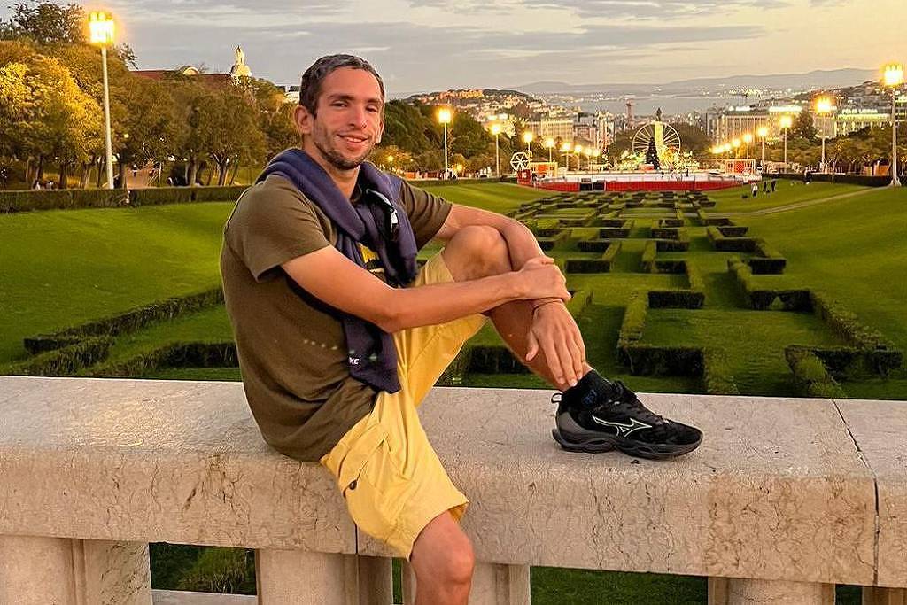 Um jovem está sentado em uma mureta de pedra em um parque. Ele usa uma camiseta verde e shorts amarelos, com um suéter amarrado ao redor do pescoço. O fundo mostra um gramado bem cuidado com arbustos podados em formas geométricas, e ao fundo há uma vista da cidade com luzes e árvores. O céu está claro, indicando o final da tarde.