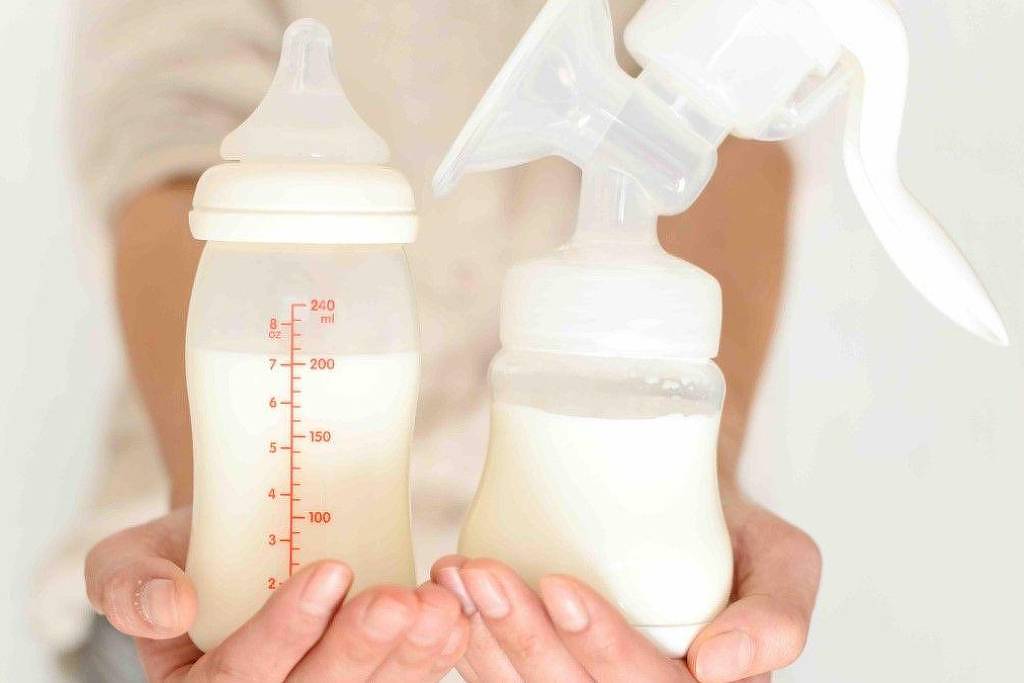 A imagem mostra uma pessoa segurando duas mamadeiras. À esquerda, uma mamadeira com escala de medição visível, contendo leite. À direita, uma bomba de leite materno, também com leite dentro. O fundo é neutro e a pessoa não está totalmente visível, apenas as mãos e os objetos que estão sendo segurados.