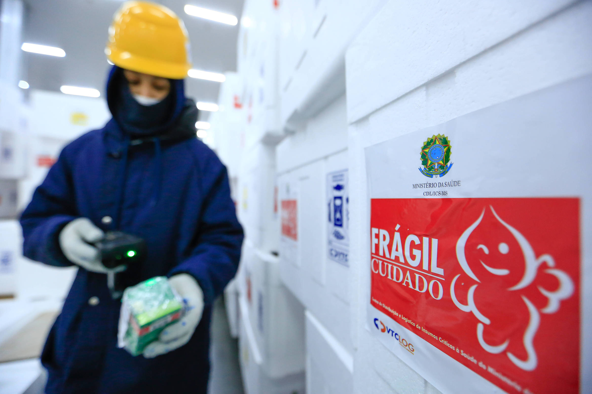 Imagem mostra uma pessoa segurando caixa de medicamento dentro do centro de distribuição de insumos do Ministério da Saúde, localizado em Guarulhos (SP). A imagem também mostra uma caixa de com adesivo escrito