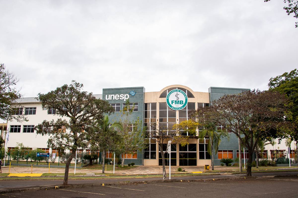 A imagem mostra a fachada de um edifício da UNESP, com o logotipo da universidade visível. O prédio é de dois andares, com janelas amplas e uma entrada central. À frente, há árvores e um espaço pavimentado. O céu está nublado.