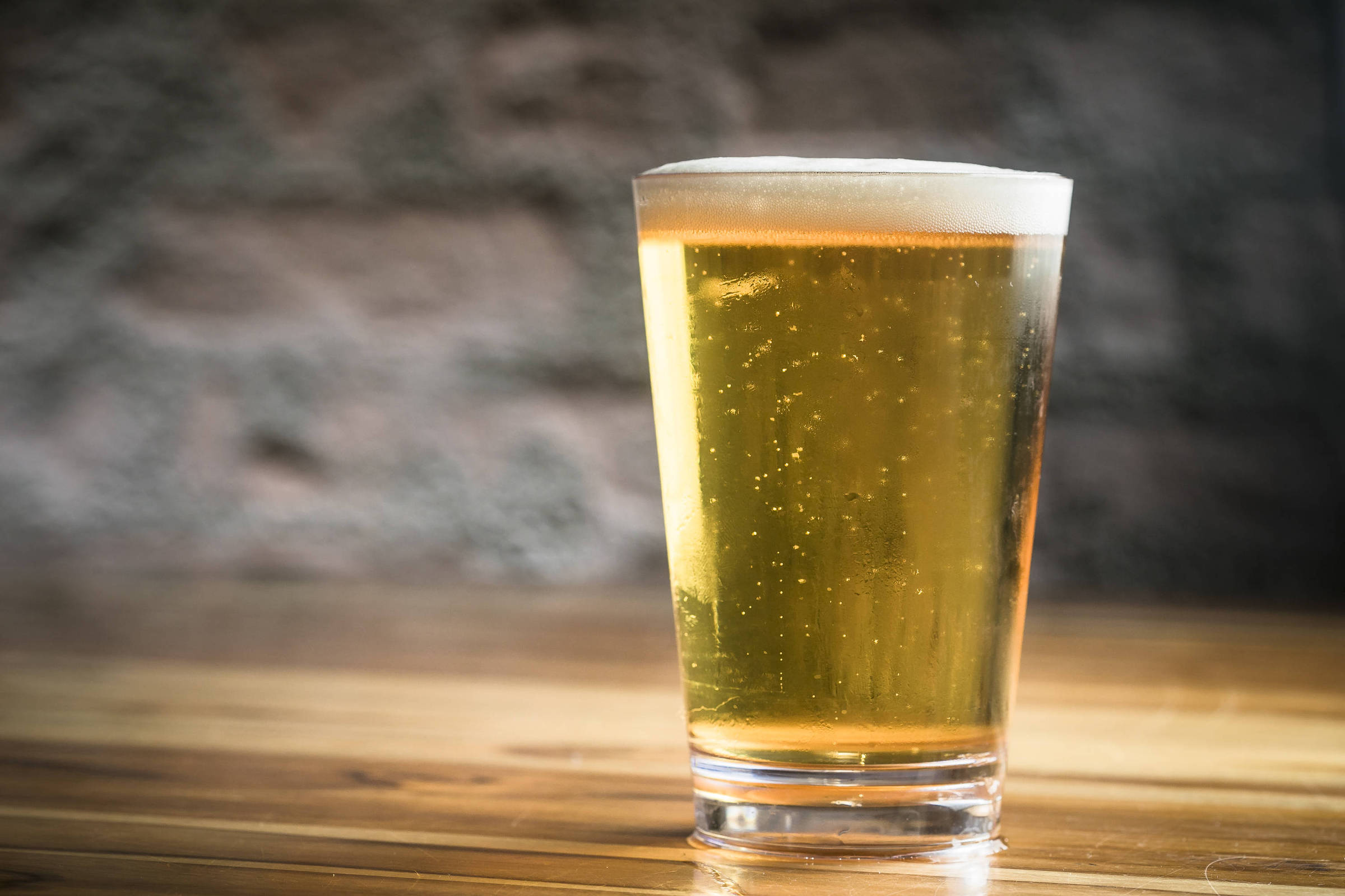 Um copo transparente de cerveja com espuma na parte superior, colocado sobre uma mesa de madeira. A cerveja é de cor dourada e apresenta bolhas visíveis, refletindo a luz ambiente. O fundo é desfocado, com uma parede de pedra ao fundo.