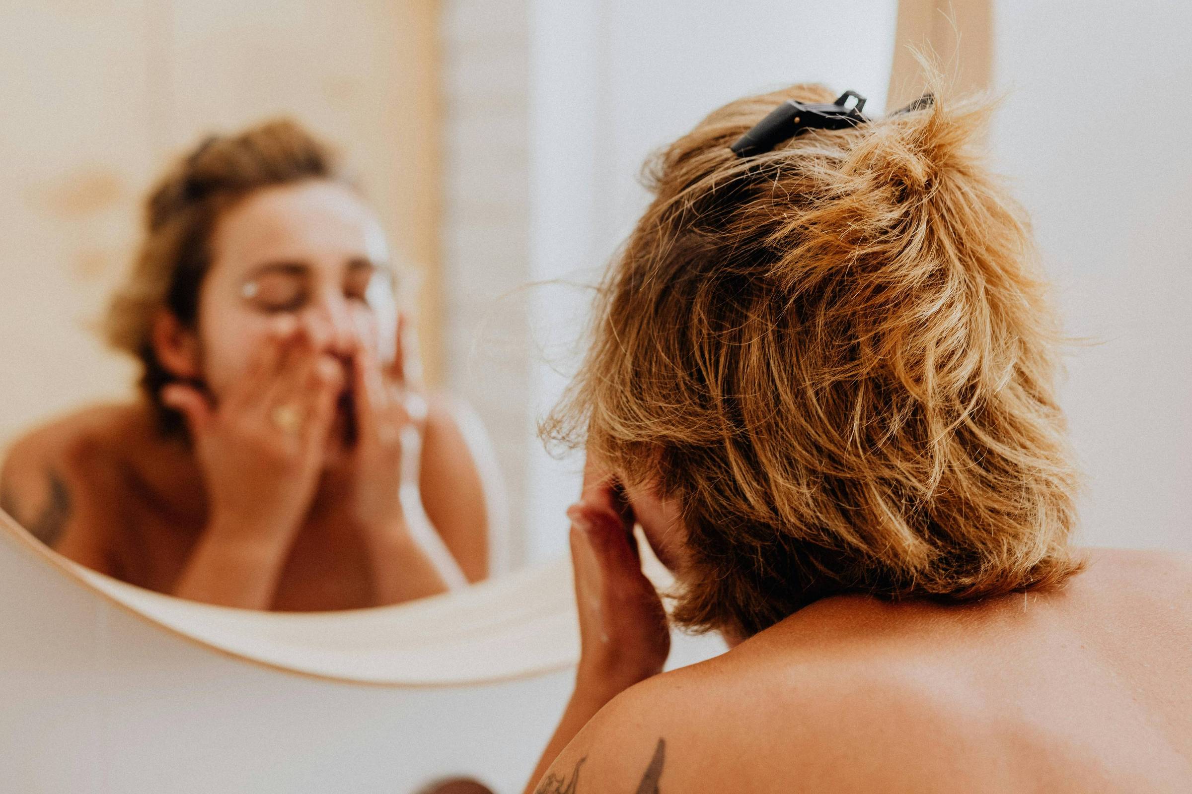 A imagem mostra uma pessoa com cabelo curto e loiro, usando uma blusa sem mangas, lavando o rosto em frente a um espelho redondo. A pessoa está com as mãos no rosto, aplicando um produto de cuidados com a pele. O fundo é claro, com uma parede de madeira ao fundo.