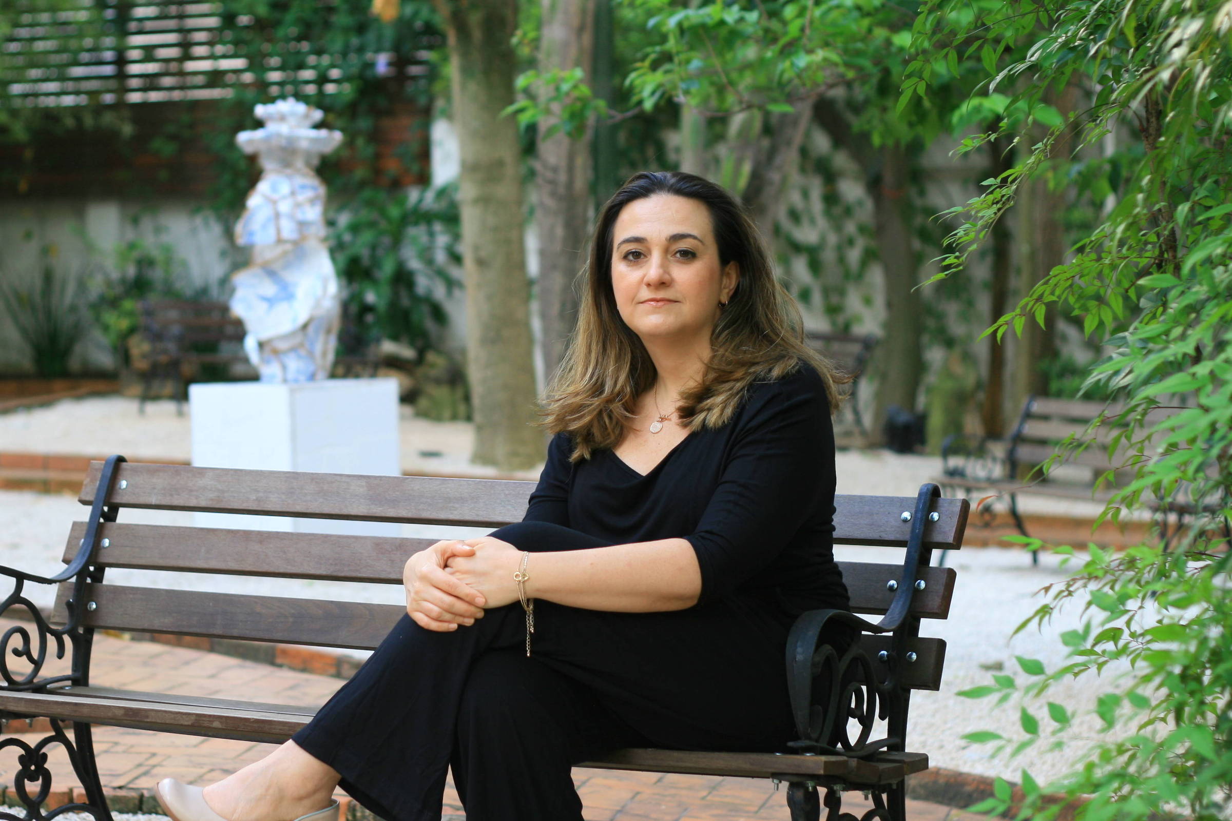 Uma mulher está sentada em um banco de jardim, vestindo uma blusa preta e calças escuras. Ela tem cabelo castanho claro e está olhando para a câmera com uma expressão neutra. Ao fundo, há árvores verdes e uma escultura branca visível, além de um caminho de pedras. O ambiente é tranquilo e bem iluminado.