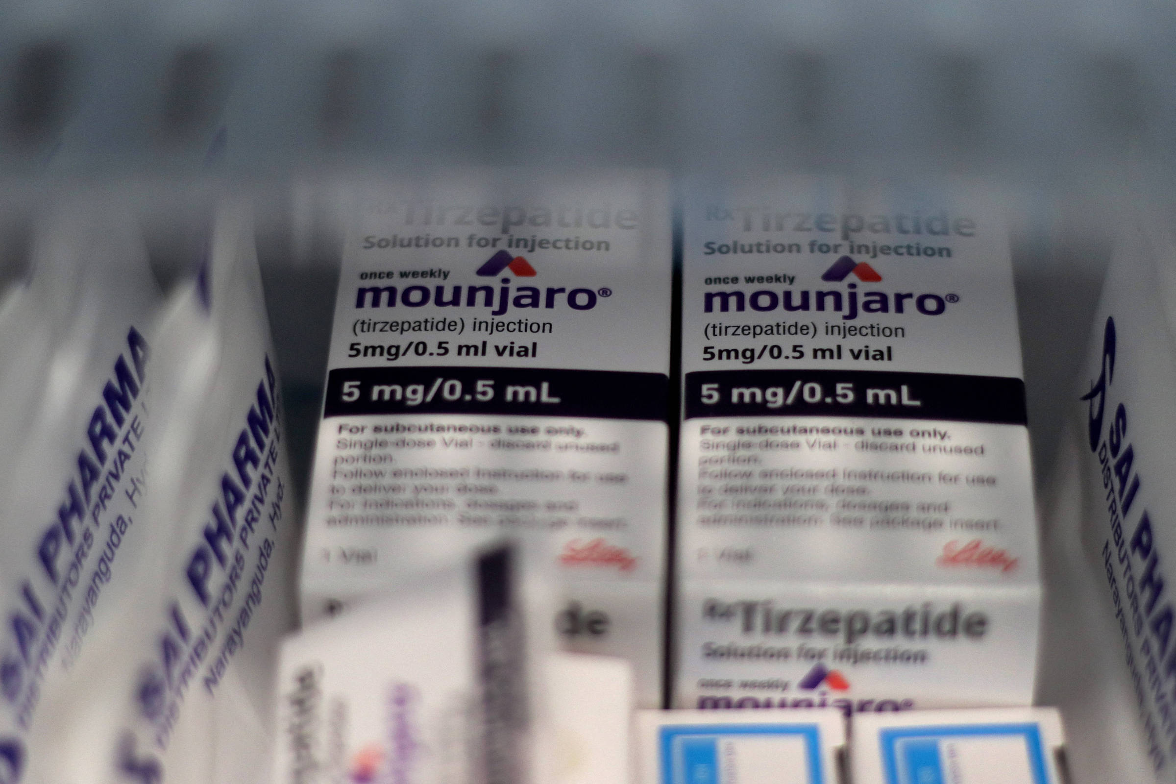 A imagem mostra frascos de medicamento Mounjaro, que contém tirzepatida, em uma prateleira. Os frascos são rotulados com a dosagem de 5 mg/0,5 mL e estão organizados em uma caixa. Ao fundo, é possível ver outros medicamentos em embalagens menores.