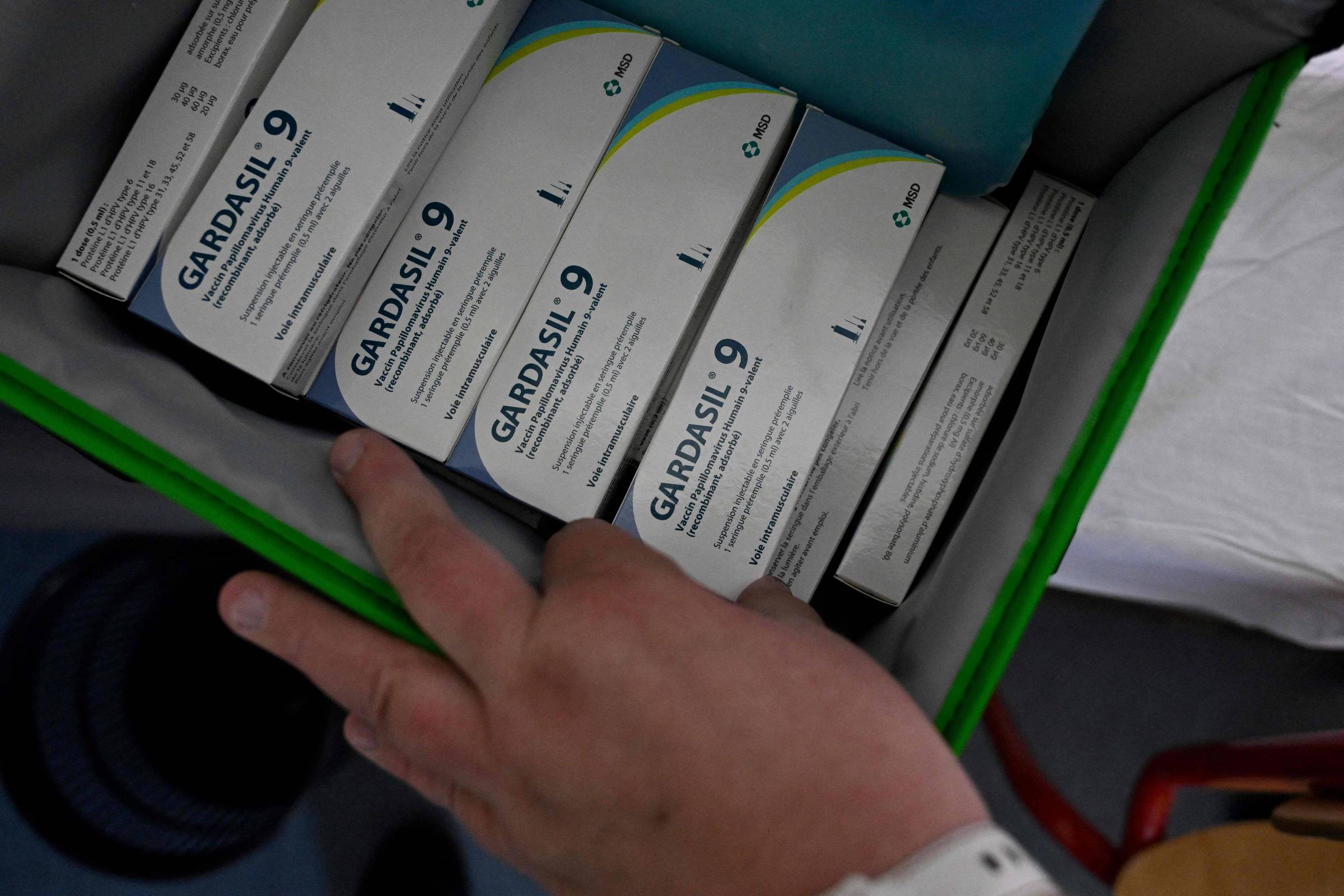 A imagem mostra uma mão apontando para caixas de vacina Gardasil 9, que estão organizadas dentro de uma bolsa ou caixa. As caixas são predominantemente brancas com detalhes em azul e verde, e contêm informações sobre a vacina. O fundo é desfocado, mas sugere um ambiente clínico.