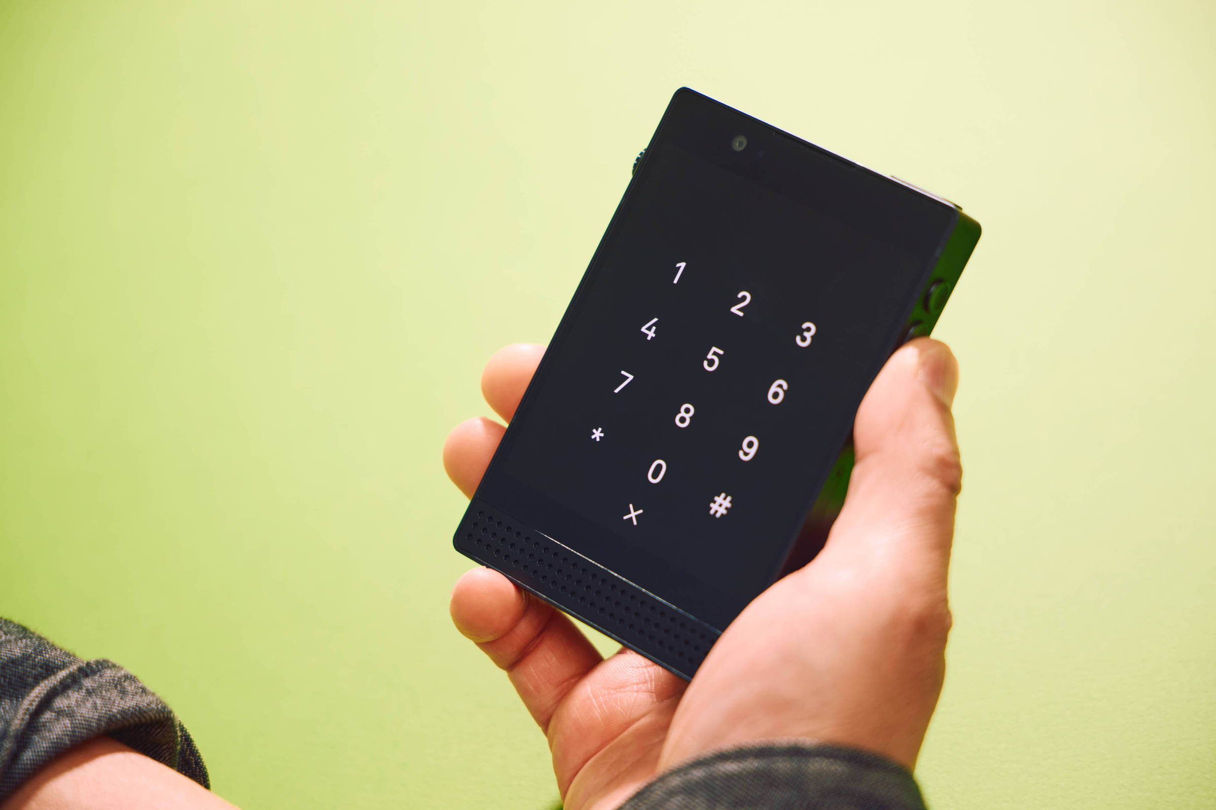 A imagem mostra uma mão segurando um celular com uma tela preta exibindo um teclado numérico. Os números de 0 a 9 estão dispostos em uma grade, juntamente com os símbolos