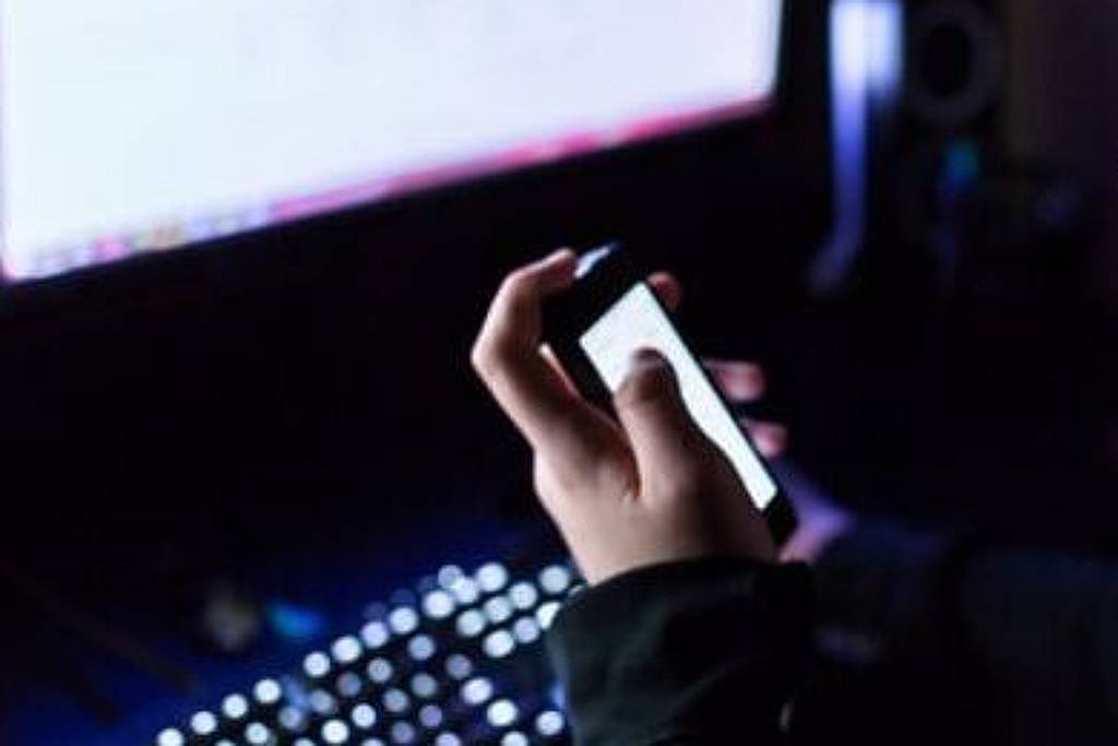 Um jovem está sentado em frente a um computador em um ambiente escuro, segurando um smartphone na mão. Ele está usando óculos e parece concentrado na tela do celular. Ao fundo, há um monitor de computador iluminado, com uma luz suave que destaca o teclado iluminado em cores. A iluminação do ambiente é baixa, criando um contraste com as luzes do teclado e da tela do computador.