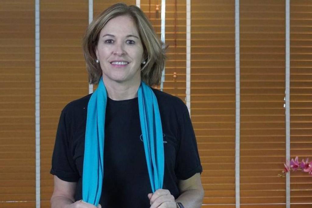 Uma mulher sorridente está em pé, usando uma camiseta preta e um lenço azul claro ao redor do pescoço. Ao fundo, há persianas de madeira e uma planta com flores rosas em uma mesa. A iluminação é suave e cria um ambiente acolhedor.