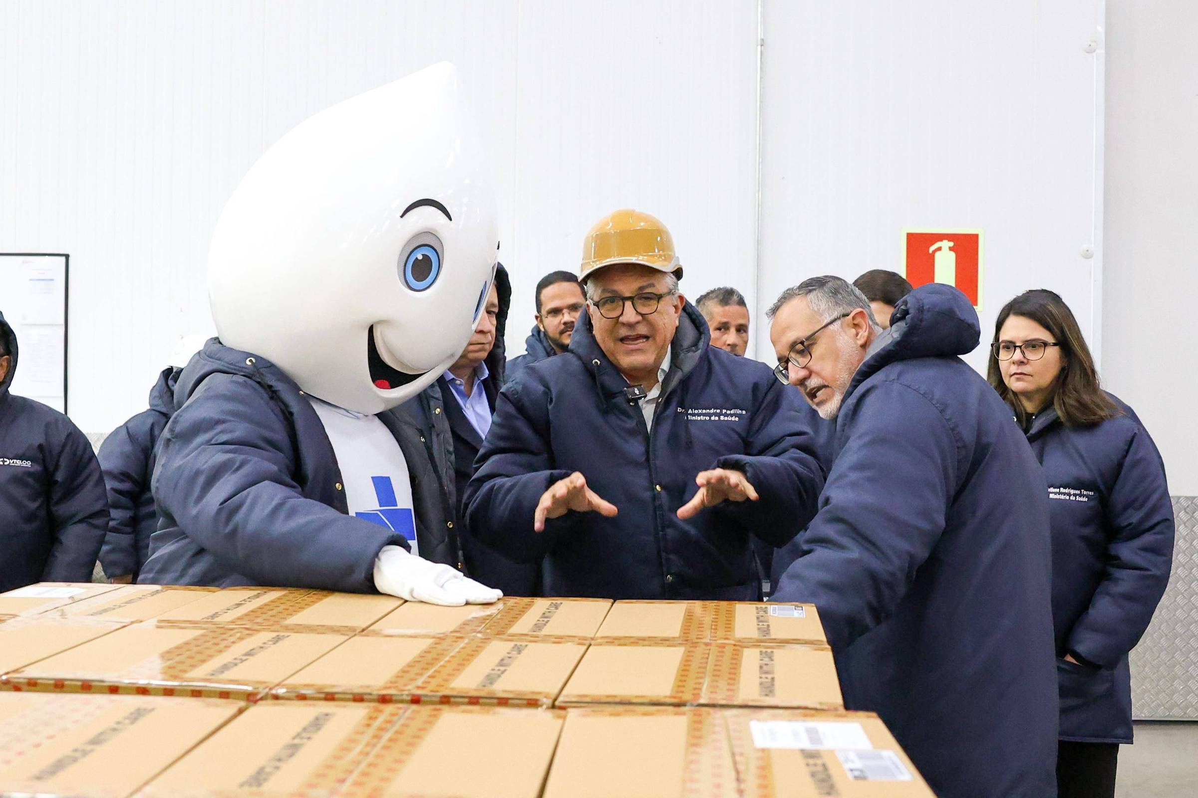 A imagem mostra um grupo de pessoas em um armazém. No centro, há um mascote com uma cabeça em forma de gota e um sorriso, vestindo um casaco azul. Ao redor do mascote, três homens estão conversando, um deles usando um capacete. Eles estão em frente a caixas empilhadas. O ambiente parece ser um local de trabalho, com paredes brancas e um sinal de emergência ao fundo.