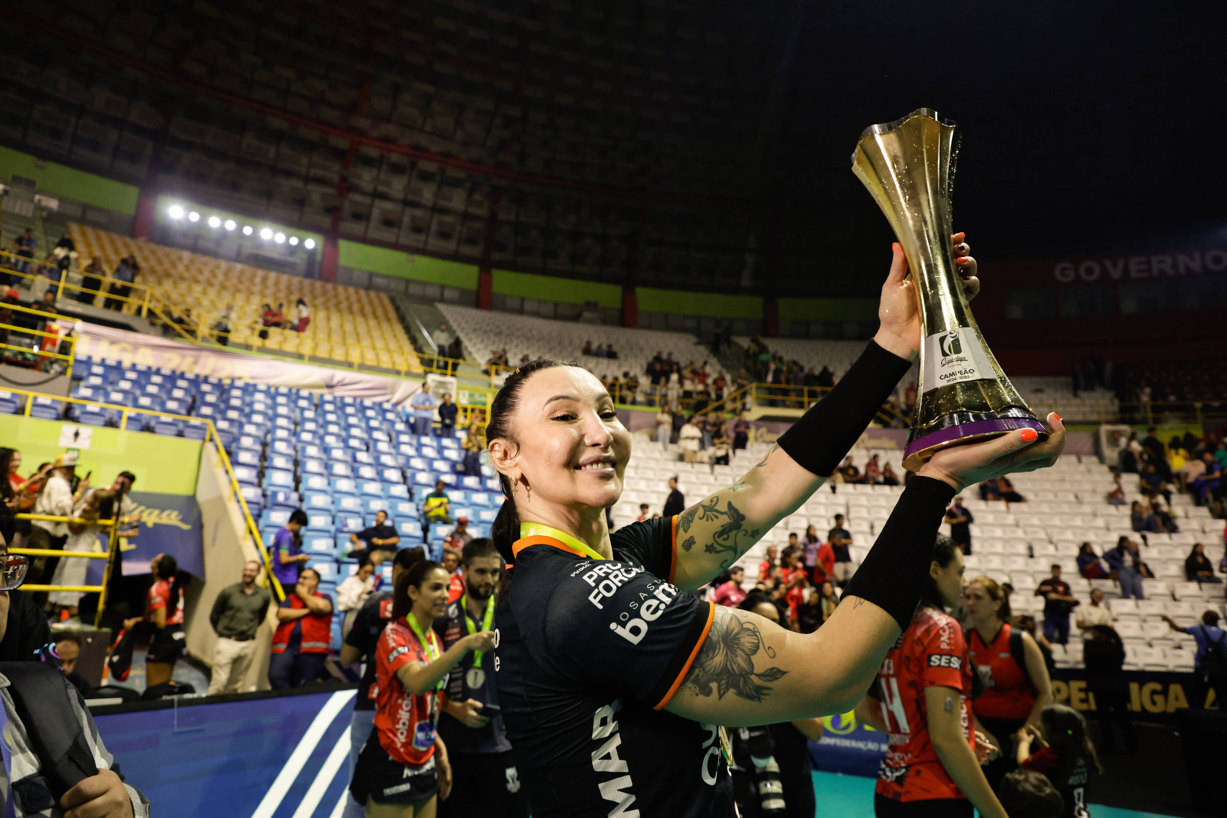 Uma jogadora de vôlei está sorrindo enquanto segura um troféu em uma quadra de vôlei. Ela está vestindo um uniforme preto com detalhes coloridos e tem tatuagens visíveis nos braços. Ao fundo, há uma plateia com algumas pessoas e cadeiras vazias, indicando que o evento pode ter acabado. A iluminação do local é intensa, destacando a jogadora e o troféu que ela ergue.
