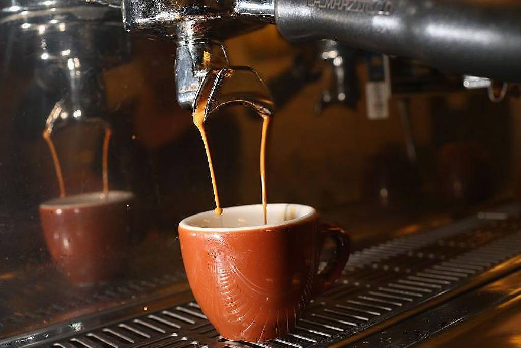 A imagem mostra uma máquina de café espresso em funcionamento, com um jato de café escorrendo de um bico para uma xícara marrom. A xícara está posicionada embaixo do bico, capturando o café que está sendo preparado. O fundo é desfocado, destacando a ação de fazer o café.