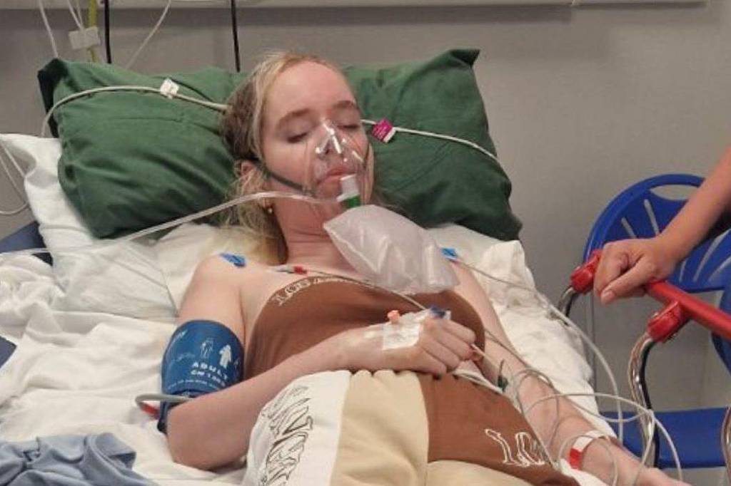 Uma jovem está deitada em uma cama de hospital, com um tubo de oxigênio no rosto e monitoramento médico visível. Ela usa um vestido de hospital e está cercada por equipamentos médicos. A jovem parece estar recebendo cuidados médicos, com um braço exposto para monitoramento e um par de mãos segurando a cama.