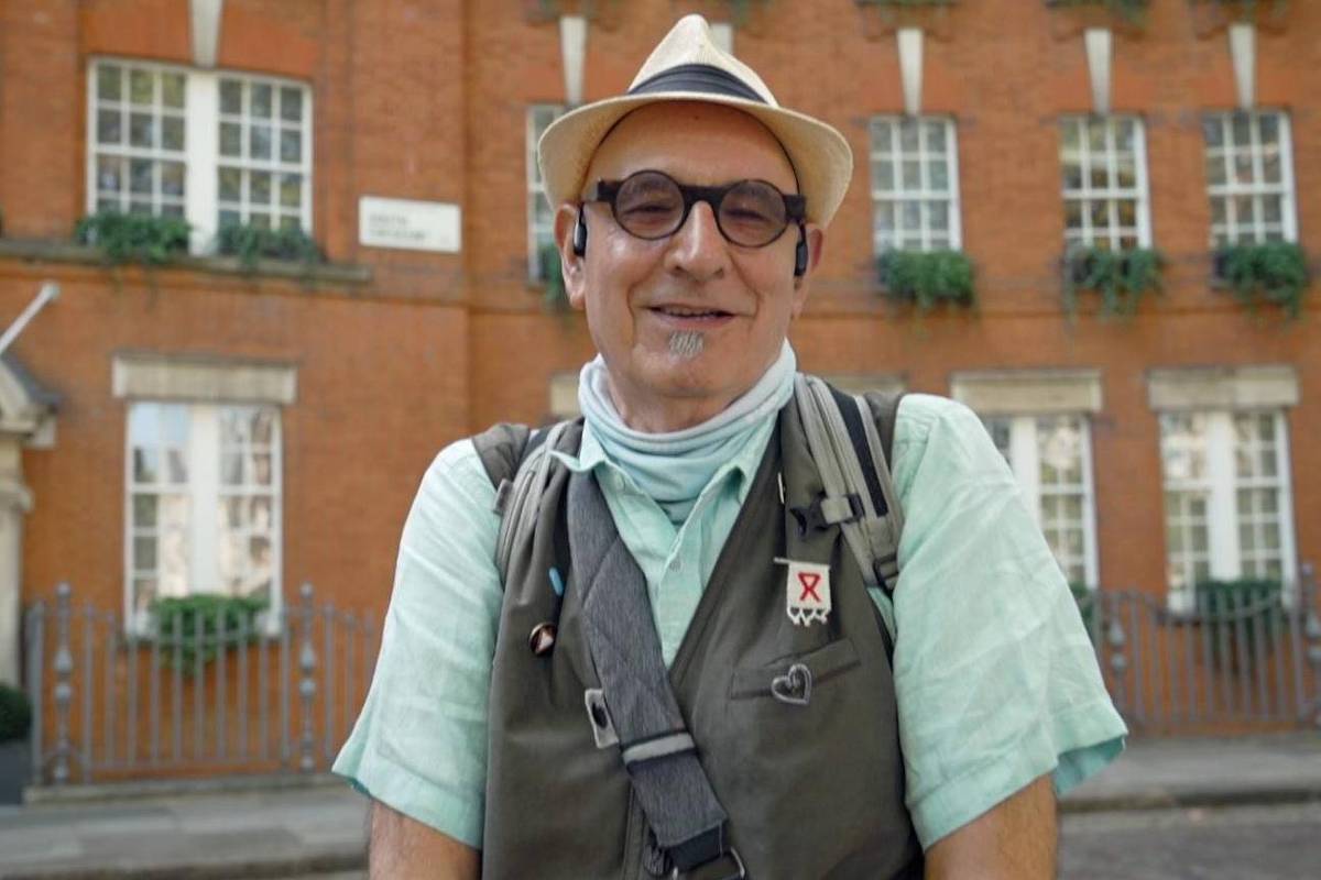 Um homem sorridente está posando para a foto em frente a um edifício de tijolos vermelhos. Ele usa um chapéu claro, óculos e uma camisa de manga curta verde. O homem também está usando um colete com vários bolsos e um lenço ao redor do pescoço. Ao fundo, há janelas grandes com plantas em vasos e uma grade de ferro na frente do edifício.