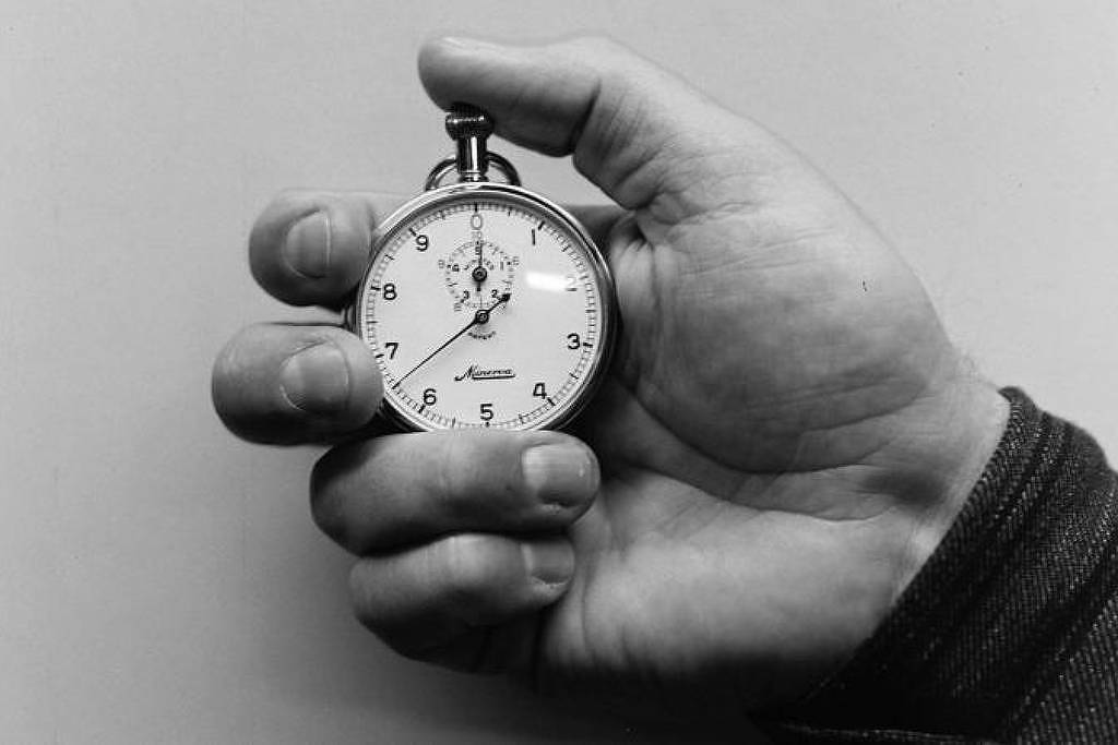A imagem mostra uma mão segurando um cronômetro prateado. O cronômetro tem um mostrador branco com números pretos e ponteiros que indicam o tempo. A mão é de um tom de pele claro e está posicionada de forma a destacar o cronômetro, que está em foco. O fundo da imagem é neutro, em tons de cinza.