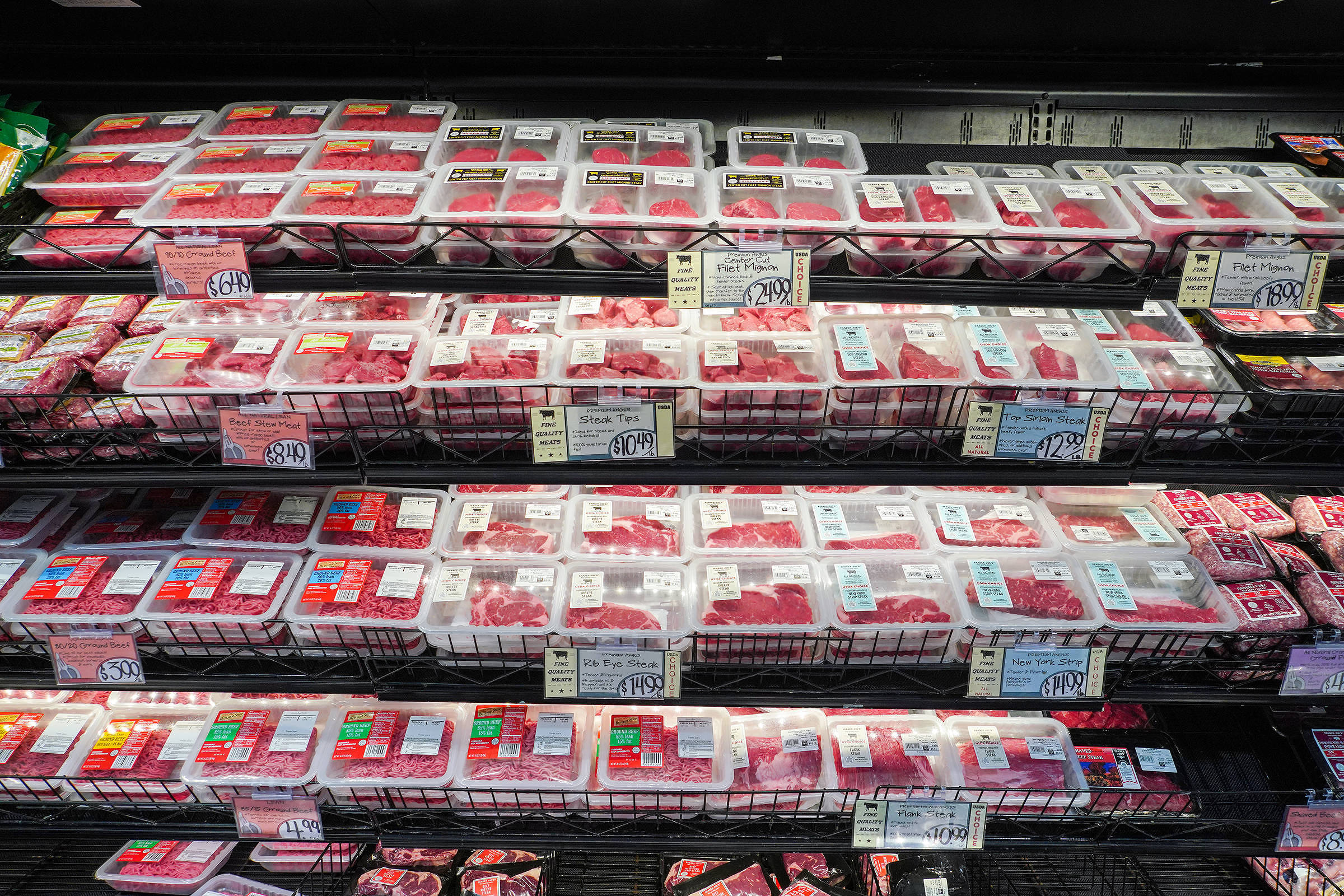 A imagem mostra uma prateleira de supermercado com várias bandejas de carne dispostas em fileiras. As bandejas contêm diferentes tipos de carne, predominantemente em tons de vermelho. Cada bandeja possui etiquetas de preço visíveis, e a prateleira está organizada em três níveis. O fundo é escuro, destacando as bandejas de carne.