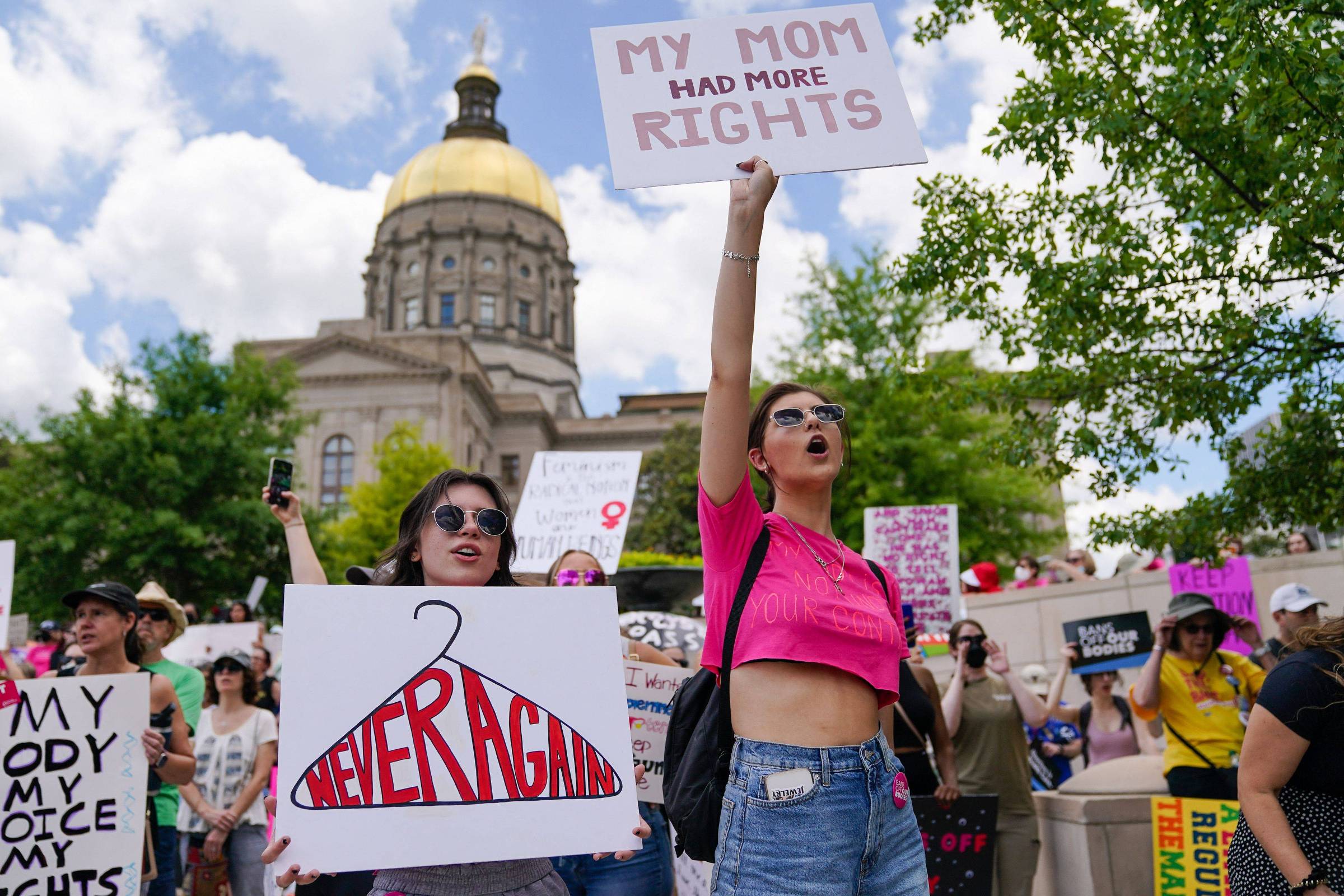 A imagem mostra um protesto ao ar livre com várias pessoas segurando cartazes. Em primeiro plano, duas mulheres estão em destaque. Uma delas, à direita, levanta um cartaz que diz