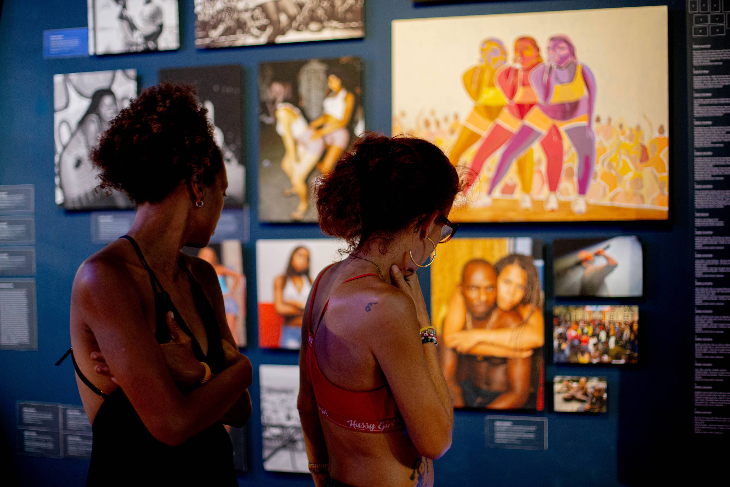 Duas mulheres estão observando obras de arte em uma exposição. Elas estão de costas, uma com cabelo cacheado e a outra com cabelo liso. As paredes ao fundo estão cobertas com várias fotografias e ilustrações, incluindo uma pintura colorida de quatro figuras femininas em trajes esportivos. A iluminação é suave, criando um ambiente contemplativo.