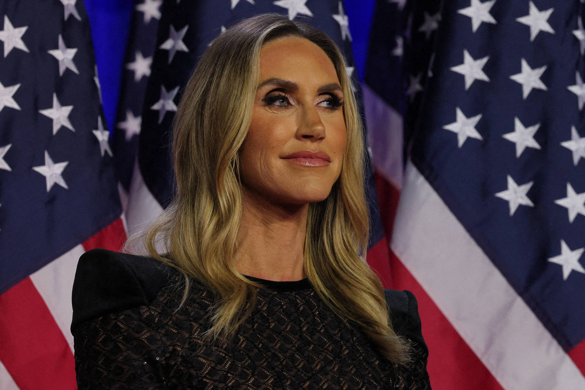 A imagem mostra uma mulher com cabelo longo e liso, usando um vestido preto com detalhes em rede. Ela está em pé, olhando para o lado, em frente a várias bandeiras dos Estados Unidos dispostas ao fundo. As bandeiras têm um padrão de listras vermelhas e brancas, com estrelas brancas em um fundo azul.