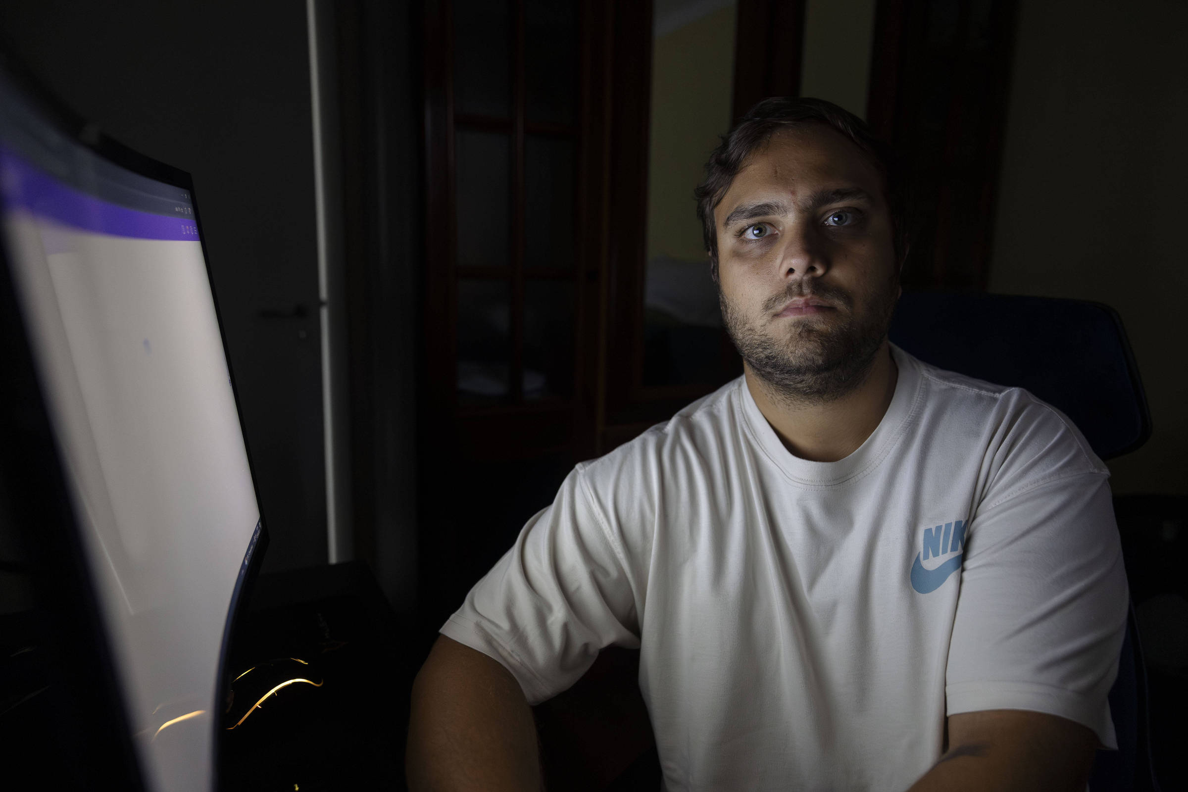 Um homem está sentado em frente a um computador em um ambiente escuro. Ele tem cabelo curto e uma barba bem aparada. O homem está usando uma camiseta branca com o logotipo da Nike em azul. A luz da tela do computador ilumina seu rosto, destacando sua expressão séria. Ao fundo, é possível ver uma porta e uma janela, mas estão em sombras devido à baixa iluminação.