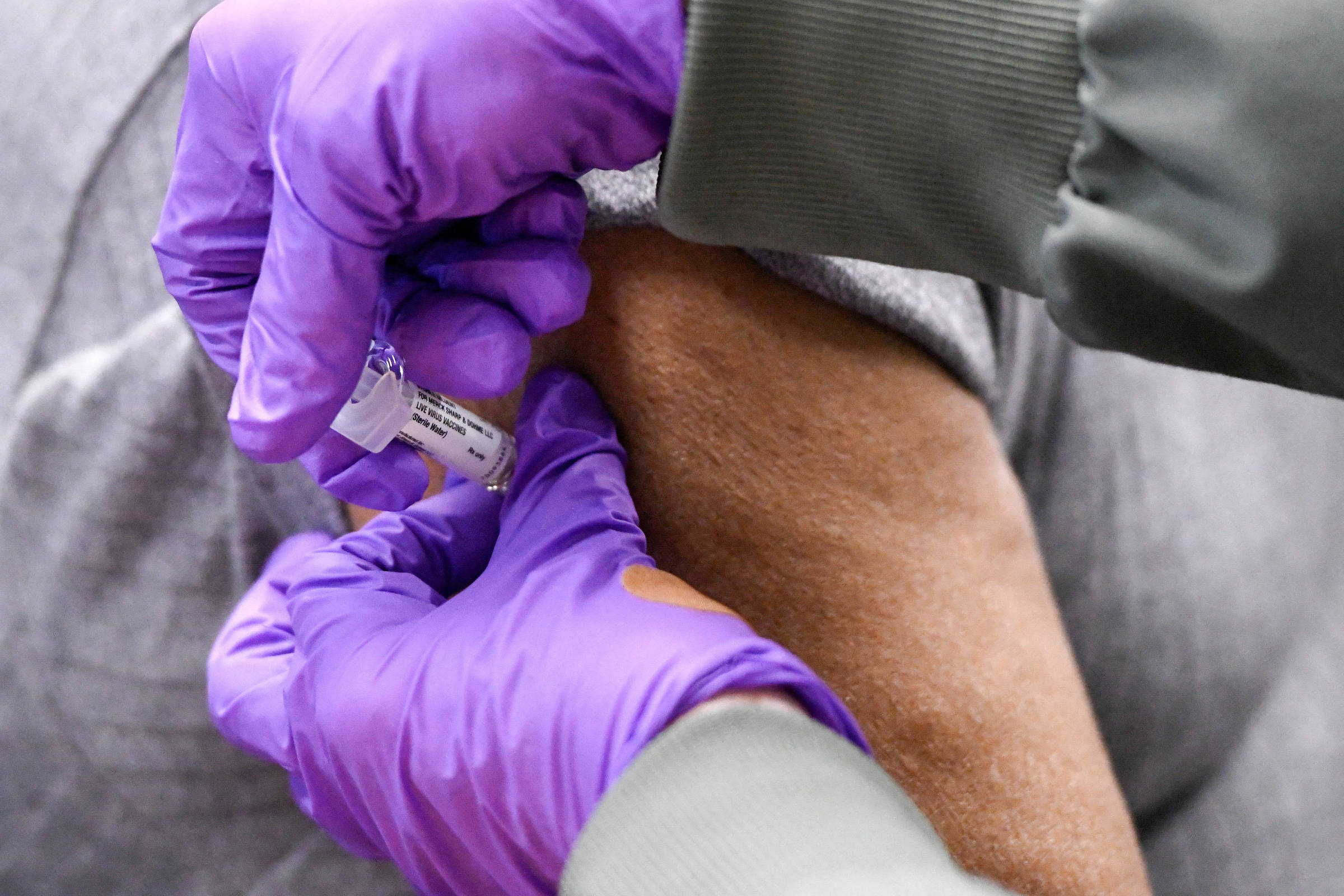 A imagem mostra uma pessoa recebendo uma vacina. Um profissional de saúde, usando luvas roxas, está segurando uma seringa com um frasco de vacina e aplicando a injeção no braço de um paciente. O braço do paciente é visível, assim como parte da roupa cinza que está sendo puxada para expor a pele.