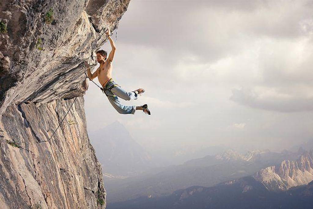 Um escalador está pendurado em uma parede de rocha vertical, usando apenas as mãos e os pés para se segurar. Ele está vestido com uma camiseta sem mangas e calças. Ao fundo, há uma vista panorâmica de montanhas e um céu nublado, criando um cenário dramático. A imagem transmite uma sensação de aventura e desafio.
