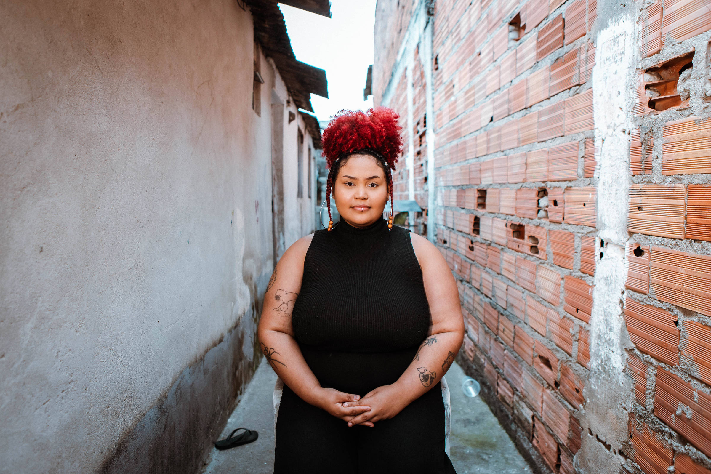 A imagem mostra uma mulher sentada em um beco estreito entre paredes de tijolos expostos. Ela tem cabelo crespo e volumoso, tingido de vermelho, e usa uma blusa preta. A mulher está olhando diretamente para a câmera, com as mãos apoiadas sobre as pernas. O ambiente ao redor é simples, com paredes de tijolos e um chão de cimento.