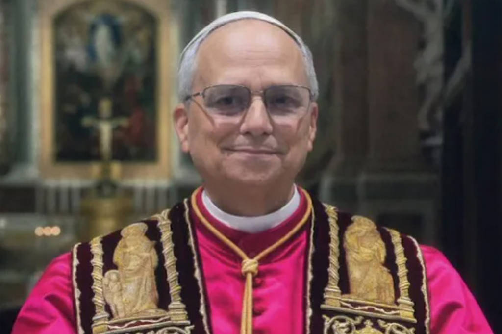 Um homem vestido com vestes papais, incluindo uma túnica rosa e uma estola branca, está posando com as mãos juntas. Ele usa óculos e tem cabelo grisalho. O fundo é uma igreja com detalhes arquitetônicos visíveis. A imagem tem uma assinatura na parte inferior que diz
