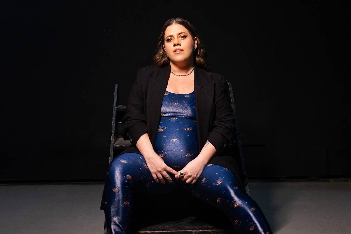 Uma mulher grávida está sentada em uma cadeira preta, com as pernas abertas e as mãos apoiadas na barriga. Ela usa um macacão azul com estampas que lembram o espaço e um blazer preto. O fundo é escuro, criando um contraste com a roupa da mulher. A iluminação é suave, destacando seu rosto e a barriga.