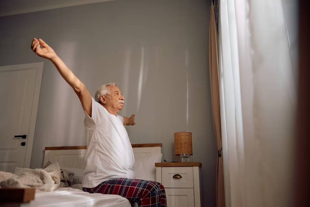 Um idoso está sentado na cama, com os braços estendidos para os lados, em um gesto de espreguiçamento. Ele usa uma camiseta branca e calças de pijama quadriculadas. O ambiente é iluminado por luz natural que entra pelas cortinas, e há uma mesa de cabeceira ao lado da cama com uma lâmpada acesa. O fundo é de uma parede clara.