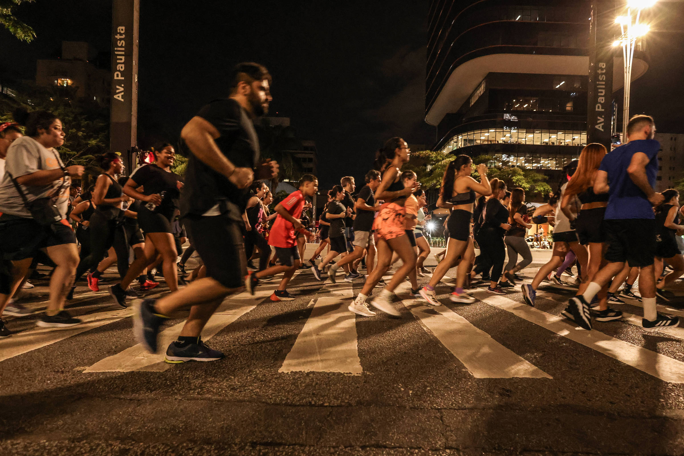 A imagem mostra um grande grupo de pessoas correndo em uma faixa de pedestres na Avenida Paulista durante a noite. As pessoas estão vestidas com roupas esportivas, algumas em tons escuros e outras em cores mais vibrantes. Ao fundo, é possível ver edifícios iluminados e árvores, criando um ambiente urbano. A iluminação da rua e dos prédios contribui para a atmosfera noturna.
