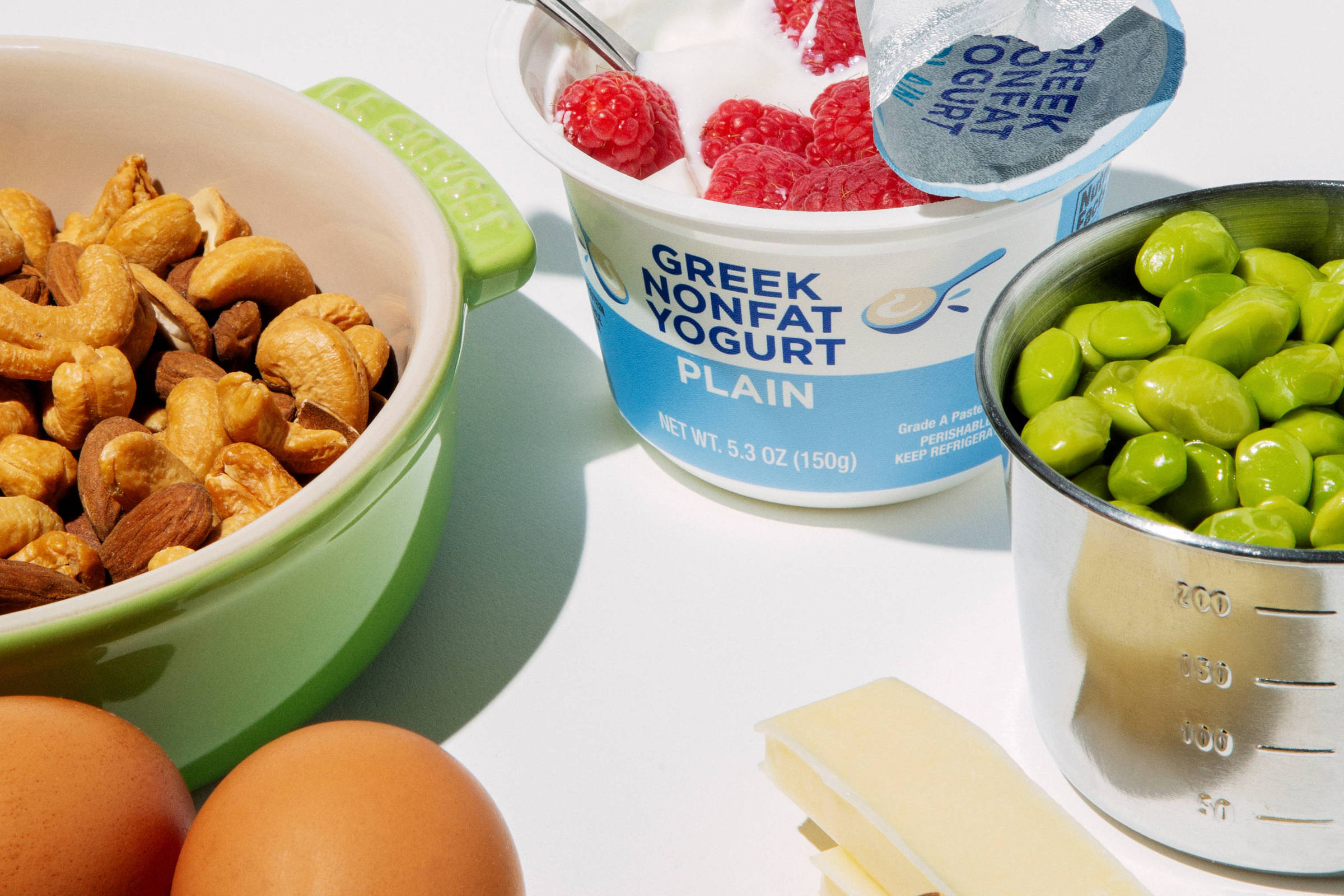 A imagem apresenta uma variedade de ingredientes saudáveis dispostos sobre uma superfície clara. Há um recipiente verde com castanhas, três ovos inteiros, um pote de iogurte grego natural com framboesas em cima, uma porção de ervilhas e um pedaço de manteiga ao lado de algumas amêndoas. Um garfo está apoiado no pote de iogurte.