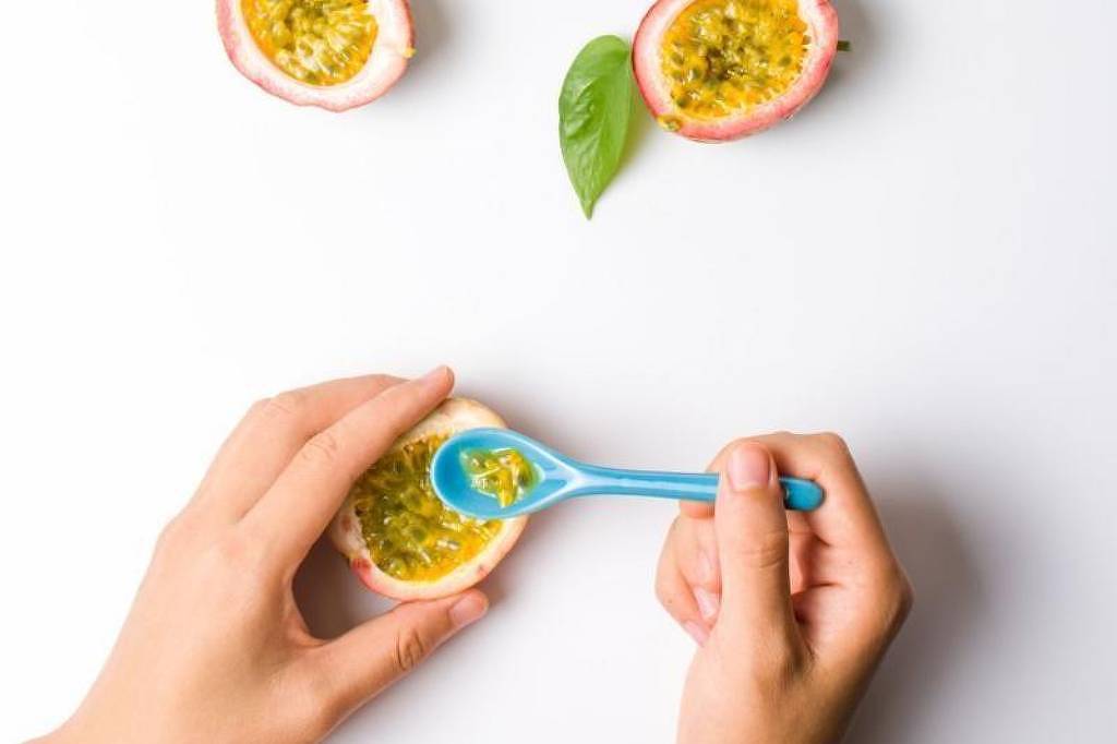 A imagem mostra várias metades de maracujá dispostas sobre uma superfície branca. Algumas metades estão acompanhadas de folhas verdes. Uma mão segurando uma colher azul está prestes a pegar a polpa de uma das metades de maracujá.