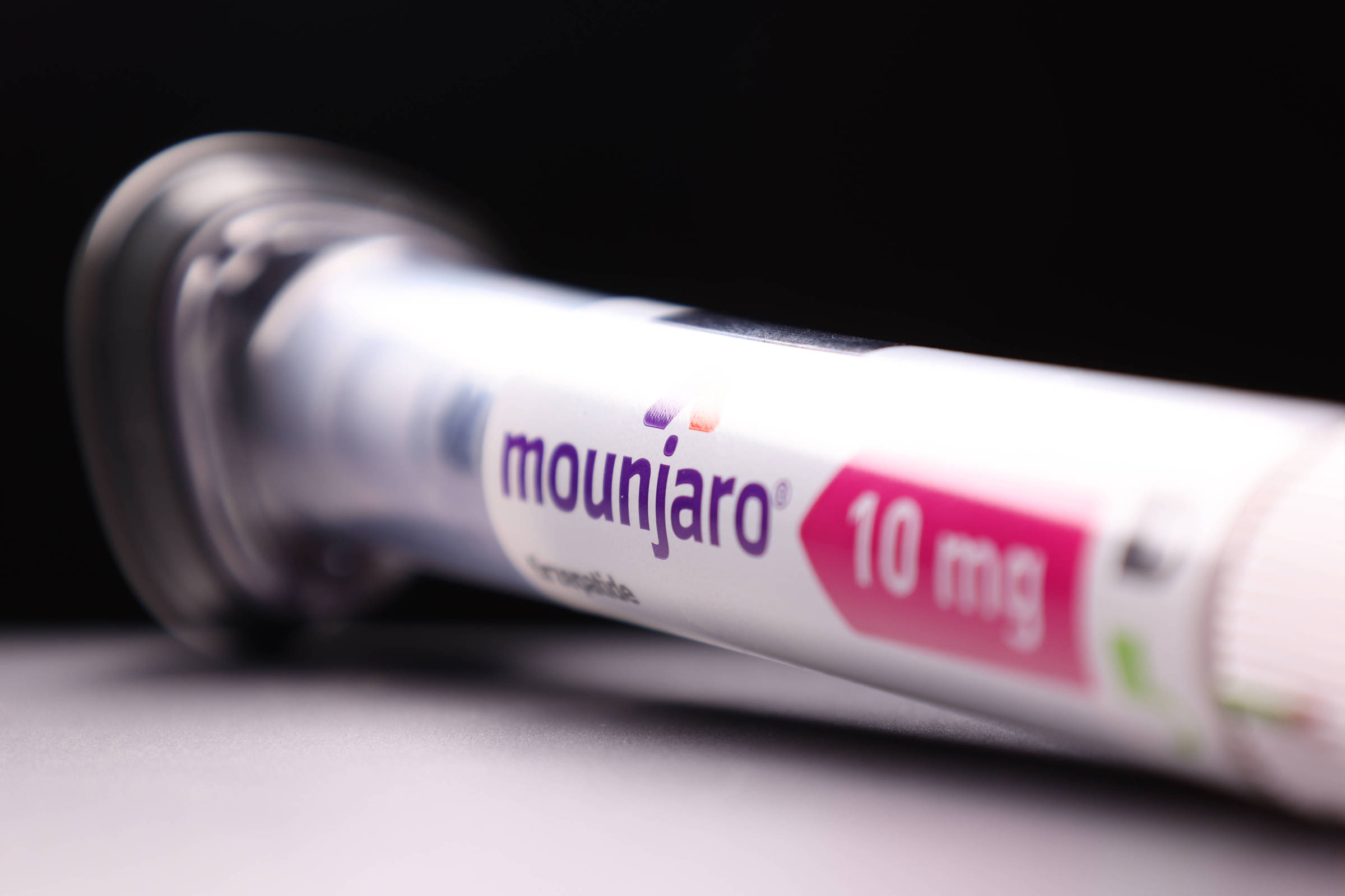 A imagem mostra uma caneta de medicamento Mounjaro, com a dosagem de 10 mg visível. A caneta está em um fundo escuro, com um design moderno e um rótulo que destaca o nome do medicamento em letras roxas e a dosagem em um fundo rosa.