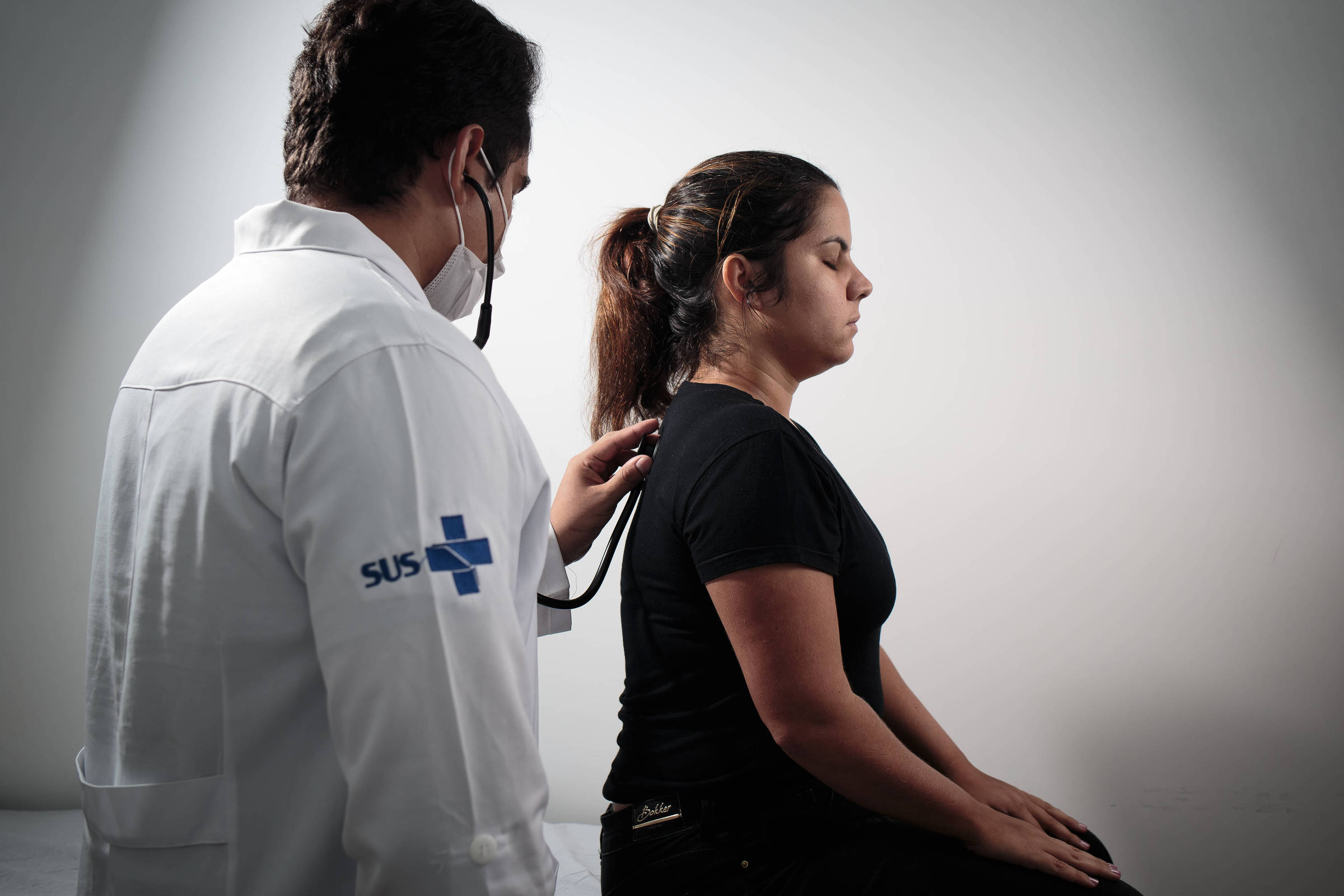 A imagem mostra um médico, vestido com um jaleco branco e usando uma máscara, auscultando uma paciente que está sentada. A paciente é uma mulher com cabelo preso, vestindo uma camiseta preta. O ambiente é neutro, com fundo claro.