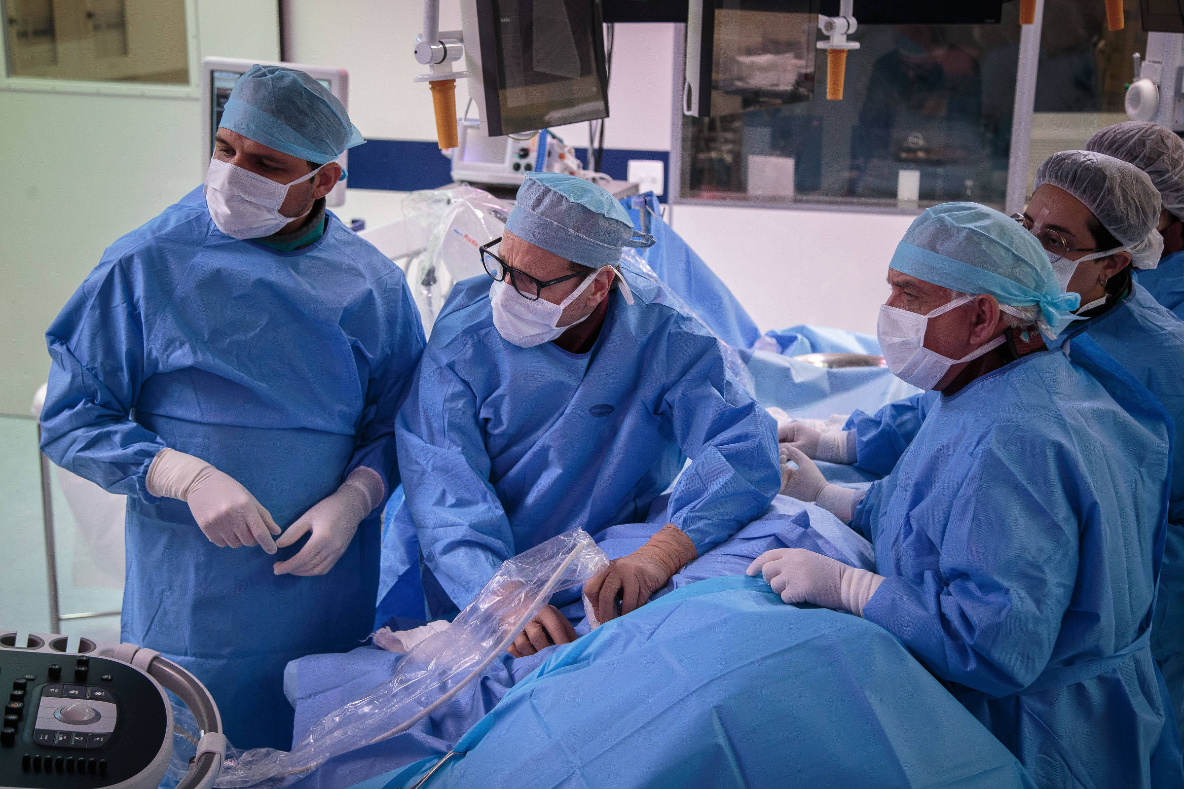 Uma equipe de cirurgiões e enfermeiros está reunida em uma sala de cirurgia, todos usando roupas cirúrgicas azuis, máscaras e toucas. Eles estão concentrados em um procedimento cirúrgico, com um paciente coberto por lençóis azuis na mesa de operação. Equipamentos médicos estão visíveis ao fundo, incluindo monitores e instrumentos cirúrgicos.