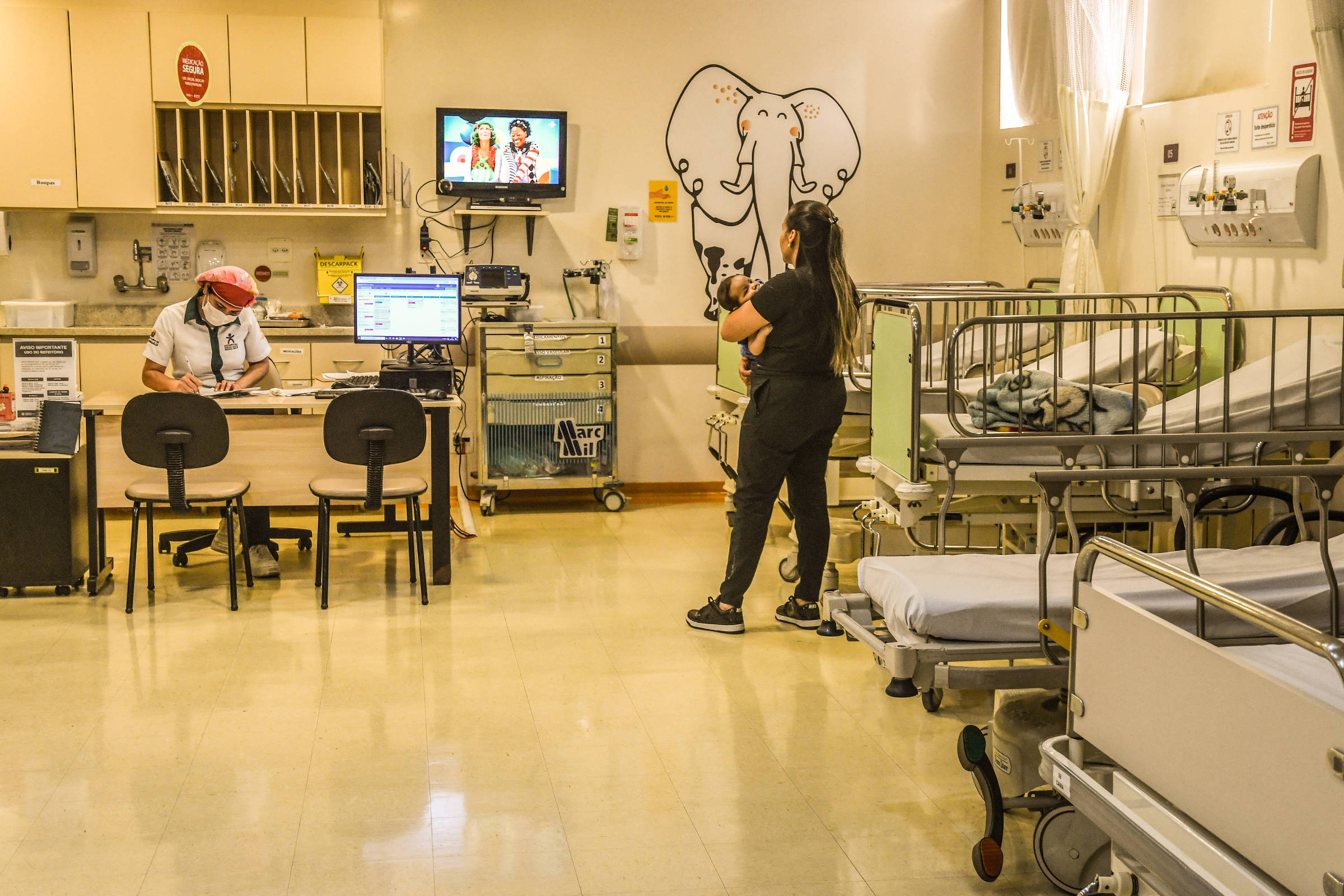 A imagem mostra o interior de um hospital, com uma recepção à esquerda onde um funcionário está sentado em uma mesa, utilizando um computador. À direita, há uma paciente em pé, vestindo roupas escuras, que parece estar conversando com alguém. Ao fundo, há várias camas hospitalares e um monitor de TV exibindo um programa infantil. A decoração inclui uma ilustração de um elefante na parede.