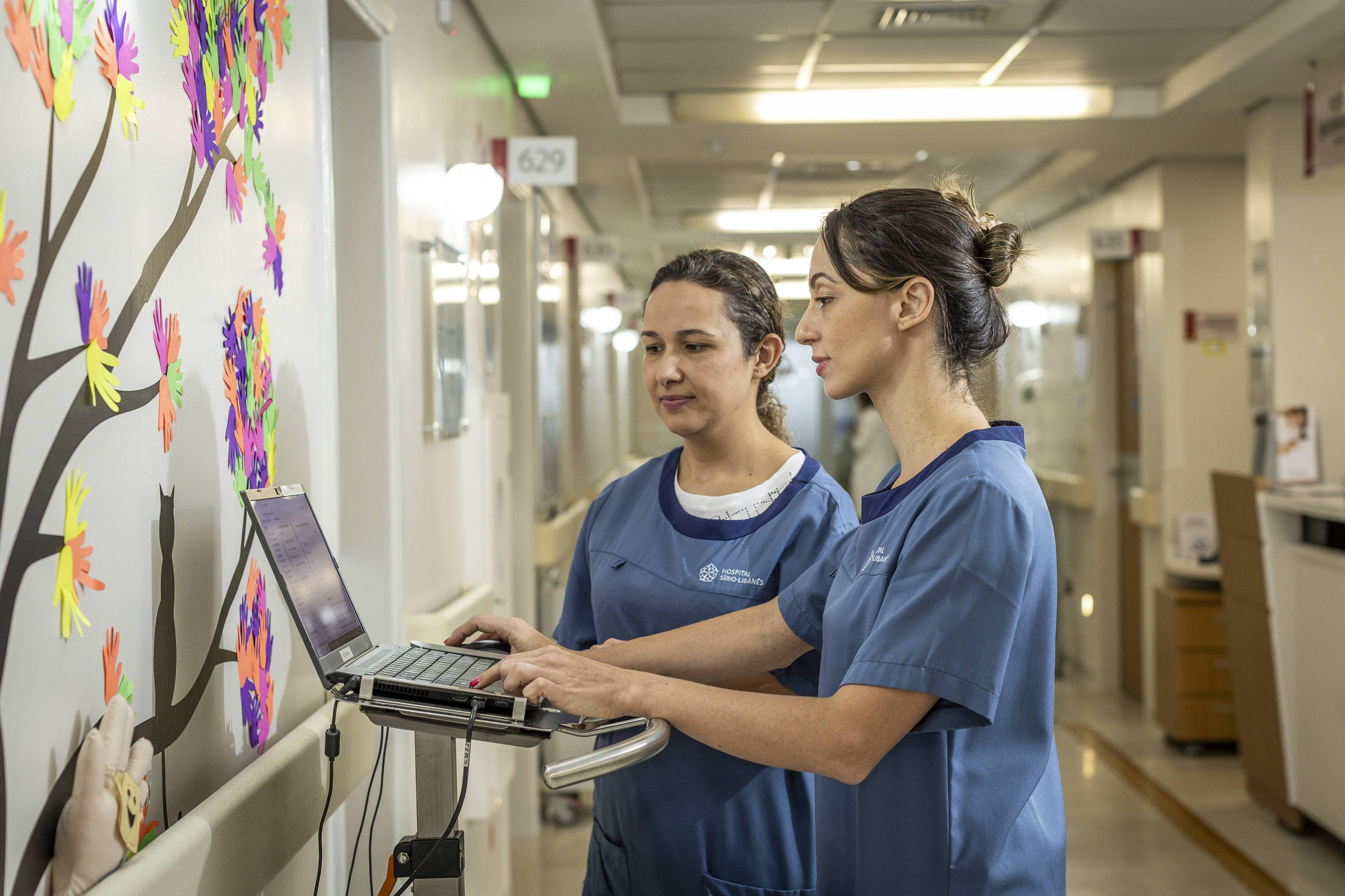 Duas profissionais de saúde, vestindo uniformes azuis, estão em um corredor de hospital. Uma delas está usando um laptop em uma mesa móvel, enquanto a outra observa. Ao fundo, há uma parede decorada com árvores e folhas coloridas, e o ambiente é bem iluminado, com portas de quartos visíveis ao longo do corredor.