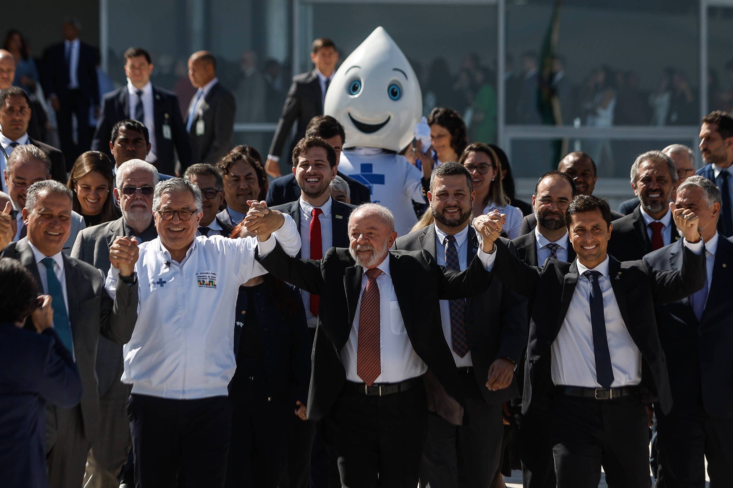 Um grupo de pessoas posando para uma foto em um evento ao ar livre. No centro, o presidente Lula, de terno escuro e gravata vermelha, segura as mãos do ministro da Saúde, Alexandre Padilha (PT). Ao fundo, está presente uma pessoa vestida de Zé Gotinha, personagem que é símbolo das campanhas de imunização do SUS.
